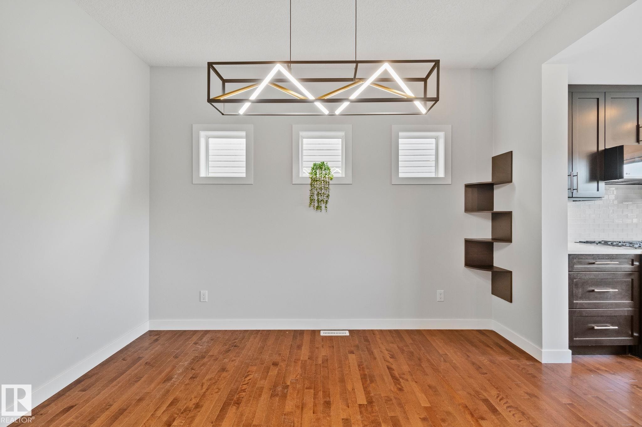 Spacious room with wood-finish flooring, white baseboards, and a contemporary linear chandelier - 614 Mcdonough Link, Edmonton, AB