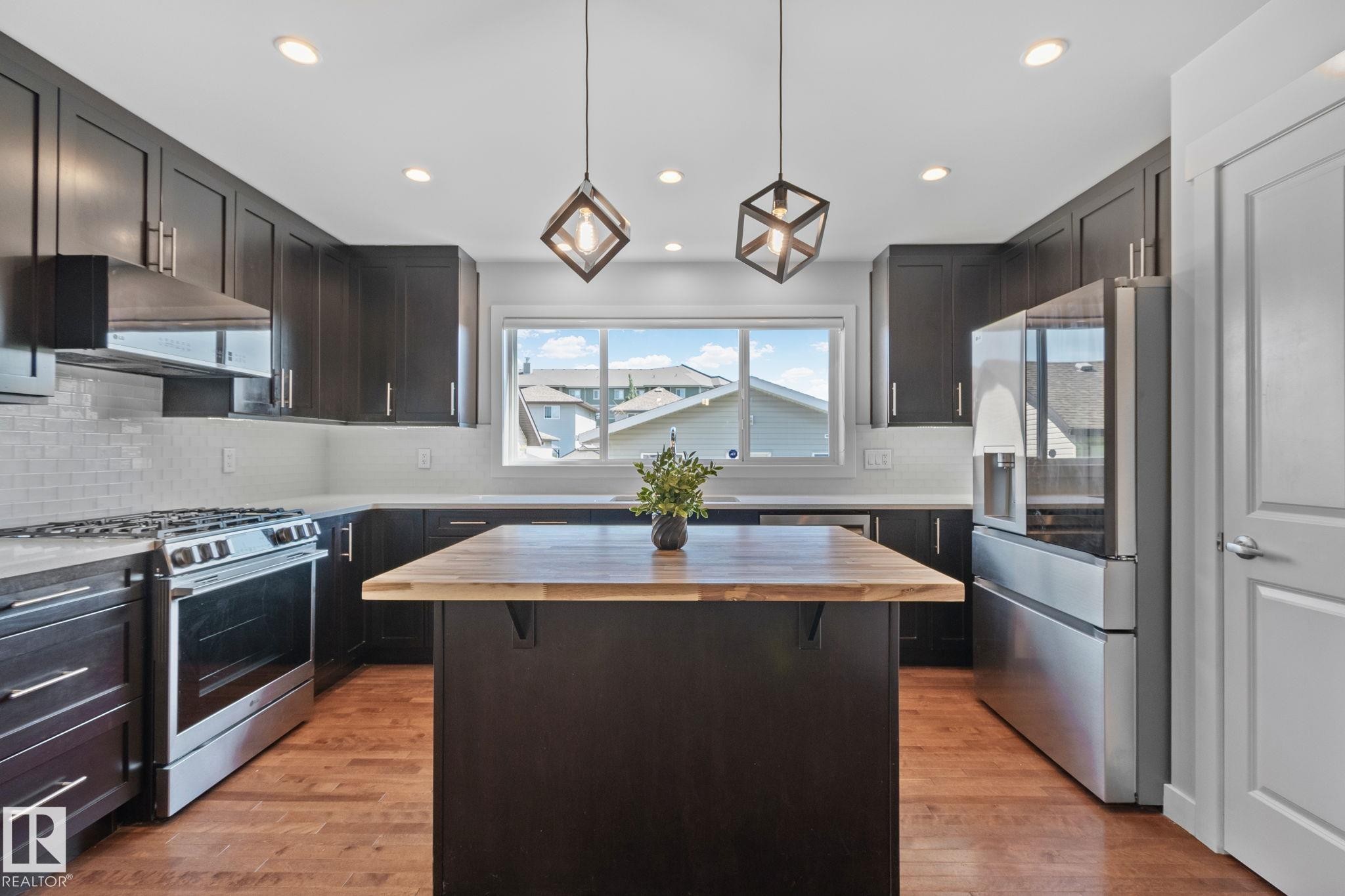 Kitchen featuring dark cabinetry, stainless steel appliances, a center island with a wood-finish countertop, white subway tile backsplash, and recessed lighting - 614 Mcdonough Link, Edmonton, AB