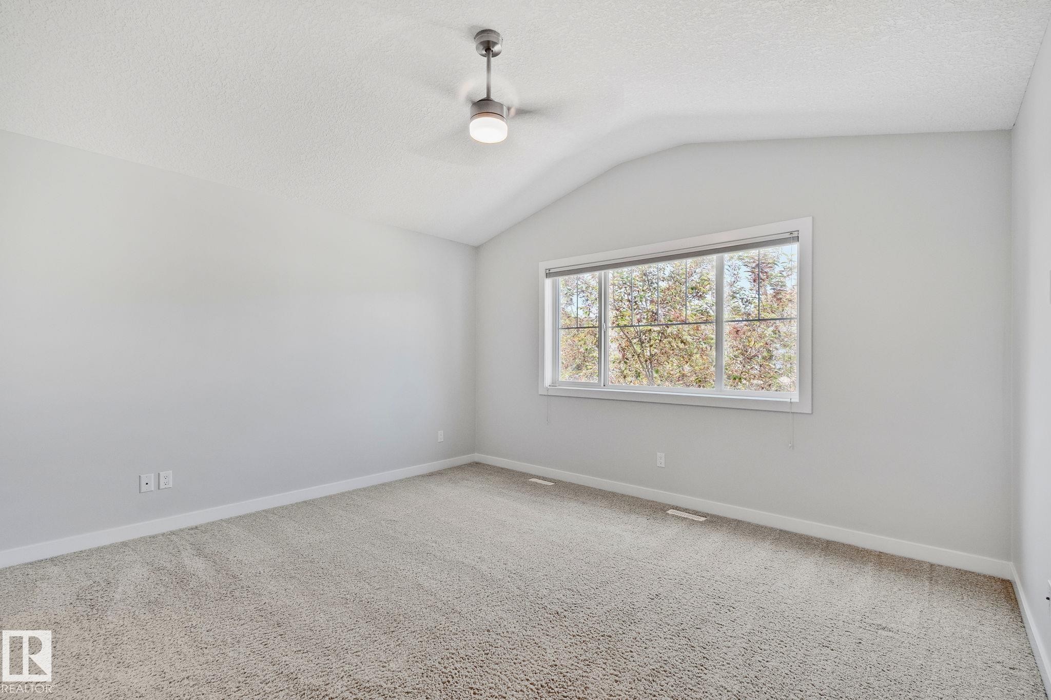 Spacious room featuring vaulted ceilings, a contemporary ceiling fan with integrated lighting, expansive window, light-colored carpeting, and neutral wall paint - 614 Mcdonough Link, Edmonton, AB