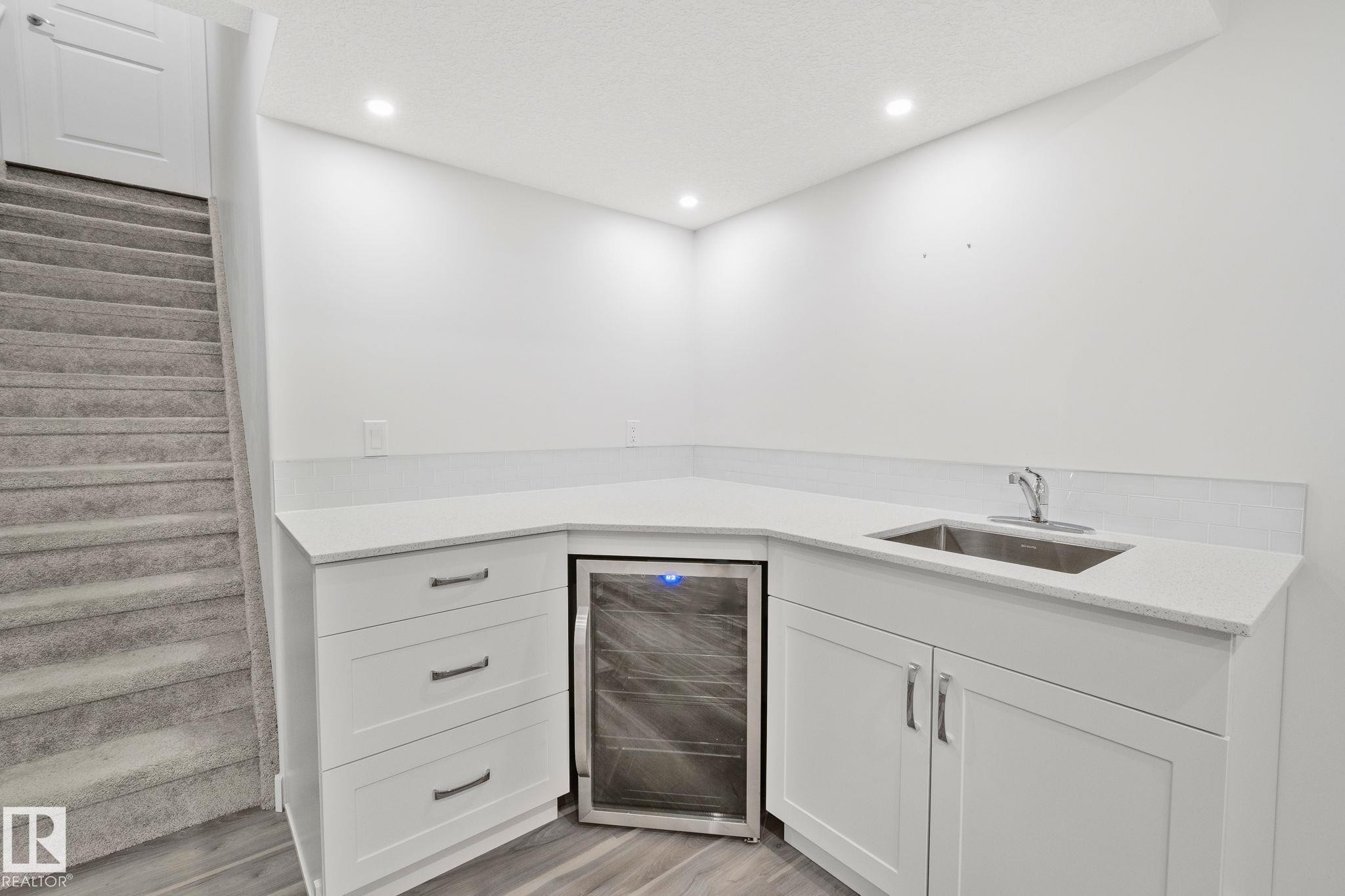 Corner wet bar featuring white cabinetry, light-toned countertops, an undermount stainless steel sink, and an integrated beverage refrigerator - 614 Mcdonough Link, Edmonton, AB