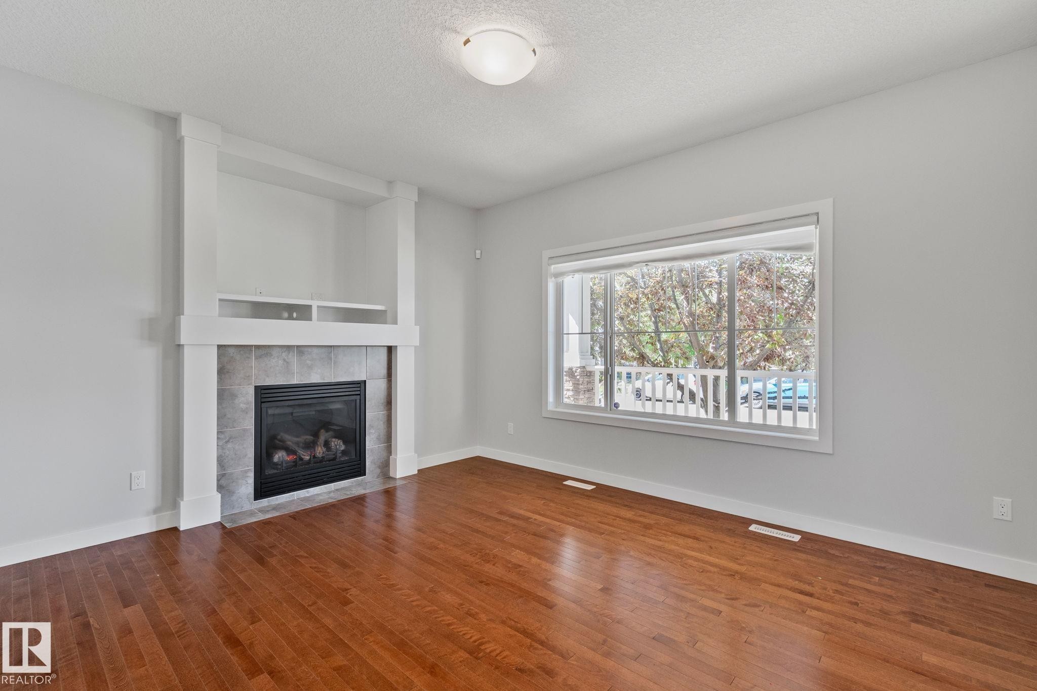 Living space featuring a tiled fireplace with an integrated media niche, rich wood-finish flooring, and a large window with white trim - 614 Mcdonough Link, Edmonton, AB