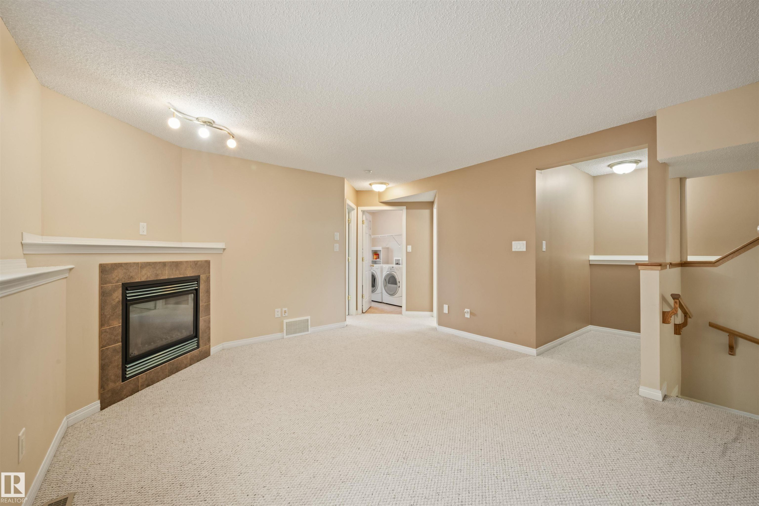 Carpeted room featuring a fireplace with a tiled surround and white mantel, track lighting, a laundry area with visible appliances, and a staircase with a wooden handrail - 7838 Schmid Place Nw, Edmonton, AB - Indoor With Fireplace