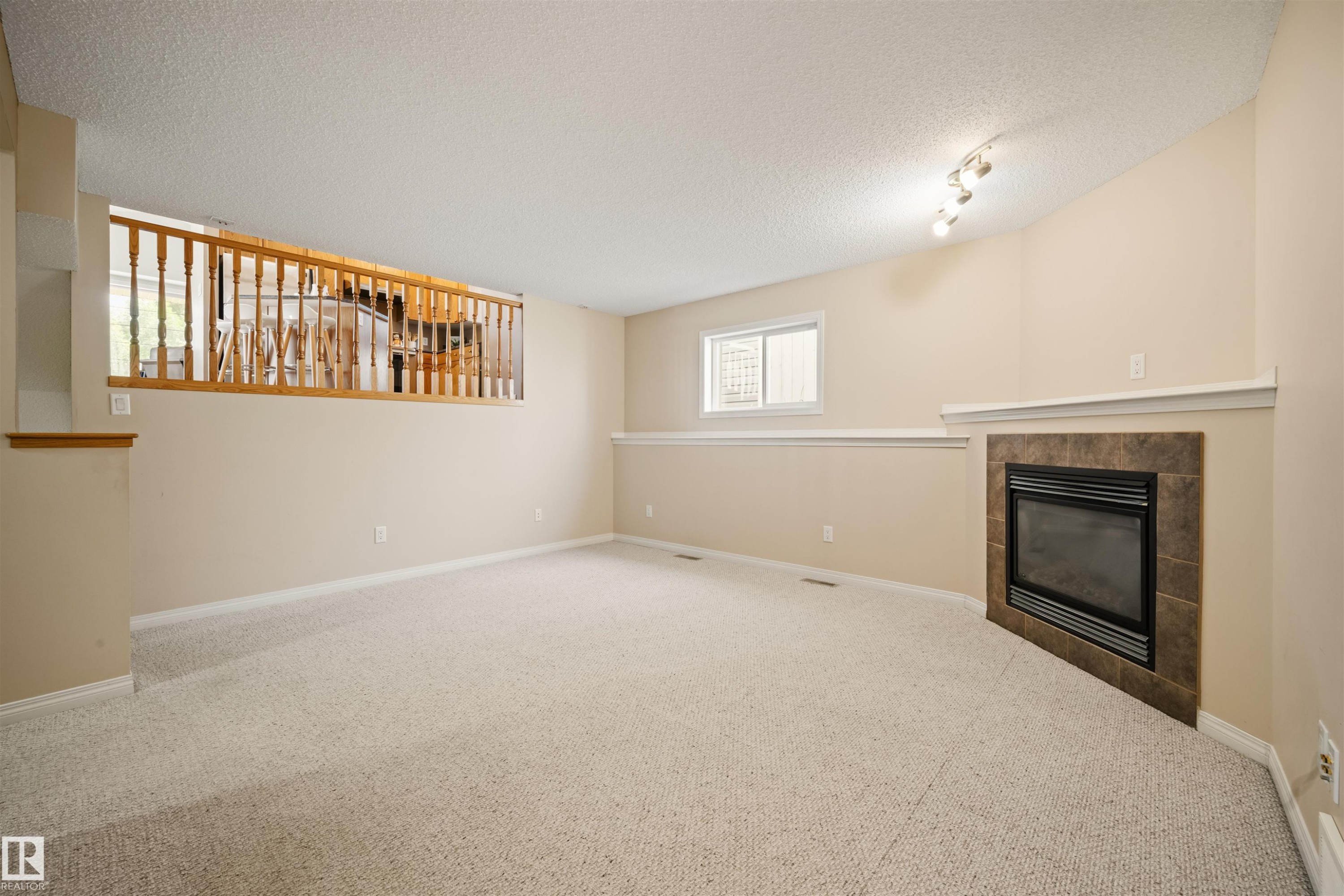 Carpeted room featuring a built-in fireplace with a tiled surround, a half-wall partition with wood railing, a window, and track lighting - 7838 Schmid Place Nw, Edmonton, AB - Indoor With Fireplace