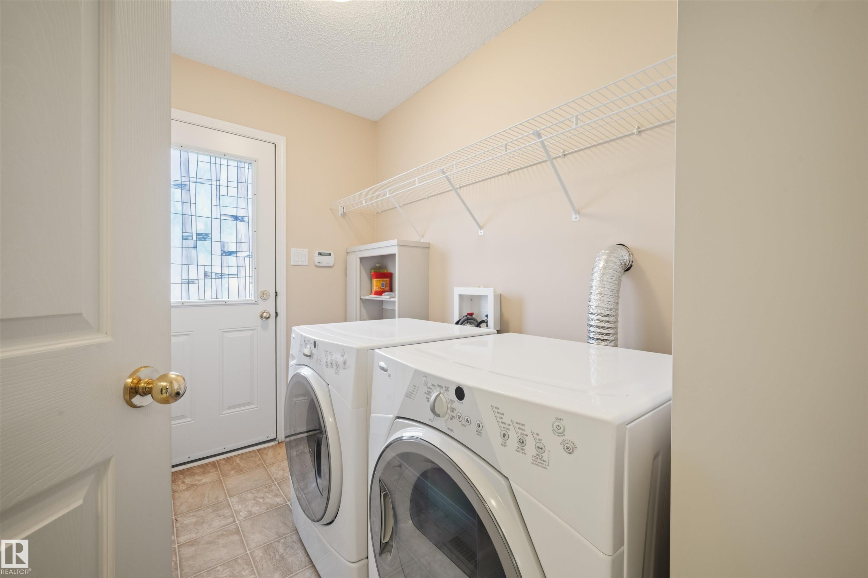 Dedicated laundry area featuring a front-loading washer and dryer, tile flooring, wire shelving, and a rear exterior door with decorative glass - 7838 Schmid Place Nw, Edmonton, AB - Indoor Photo Showing Laundry Room