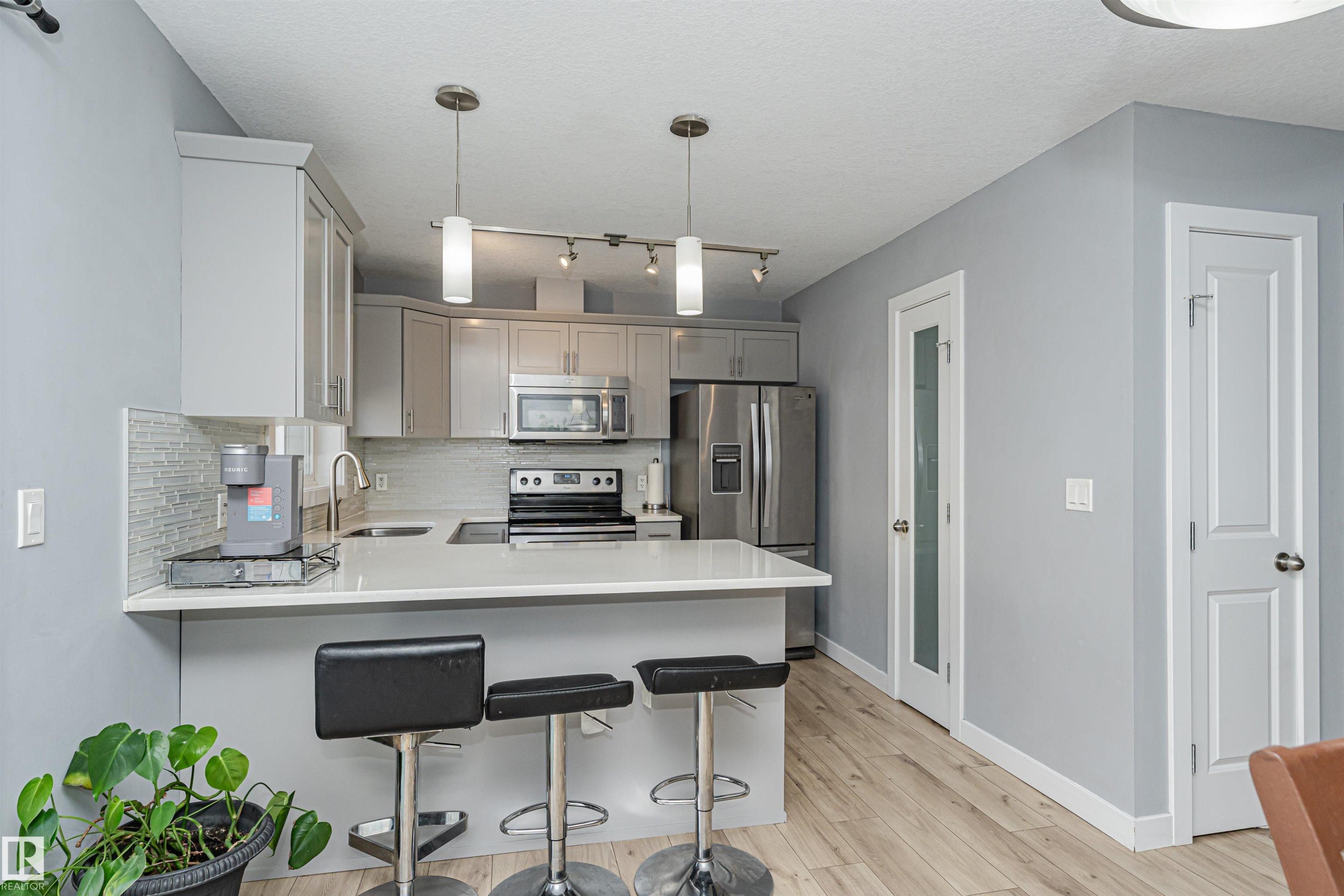 Contemporary kitchen featuring light gray cabinetry, stainless steel appliances, a tile backsplash, light wood-finish flooring, and a breakfast bar with pendant lighting - 35 675 Albany Way, Edmonton, AB