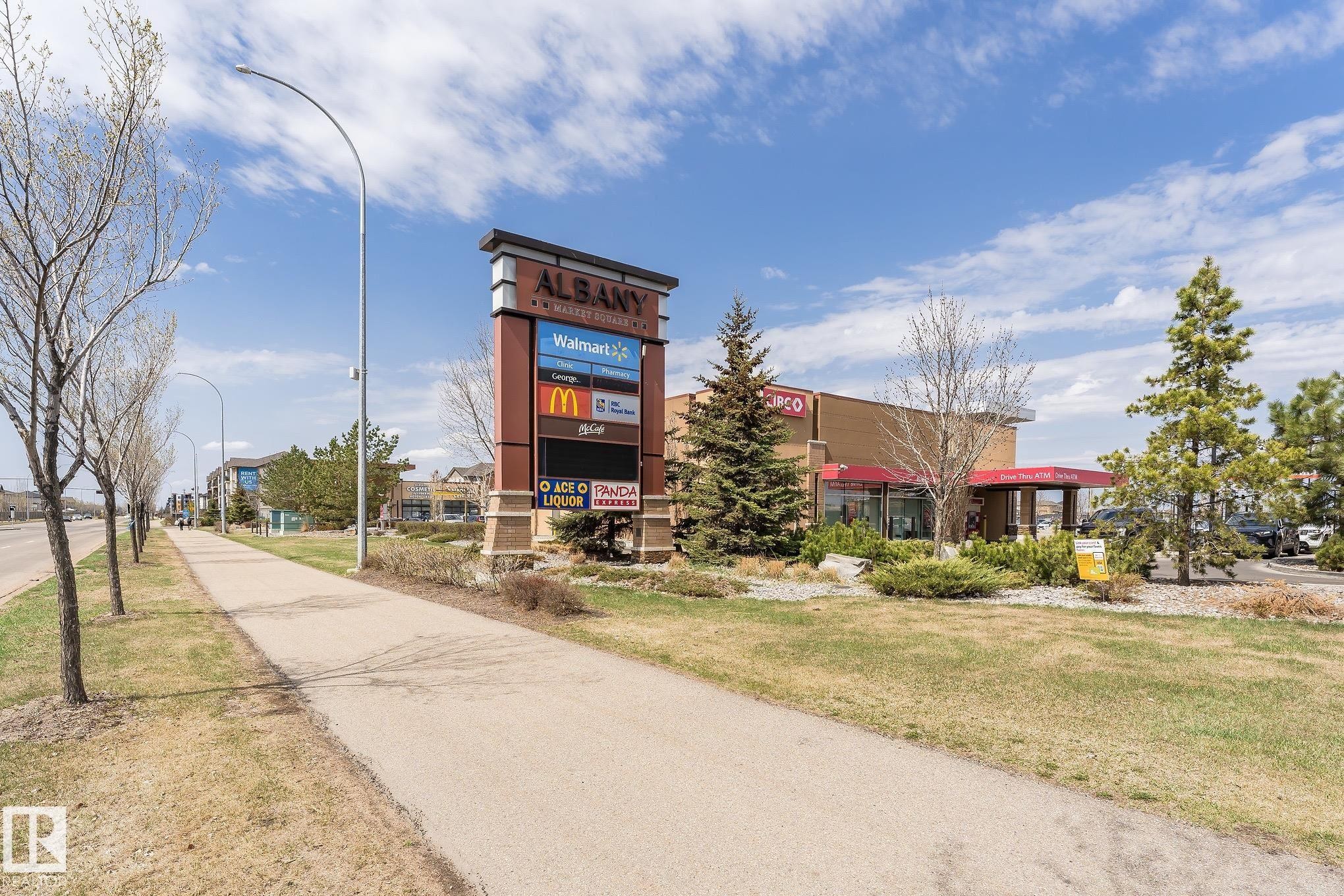 Commercial plaza with a prominent tenant directory sign, established landscaping including mature evergreen and deciduous trees, and a commercial building featuring a red awning - 35 675 Albany Way, Edmonton, AB