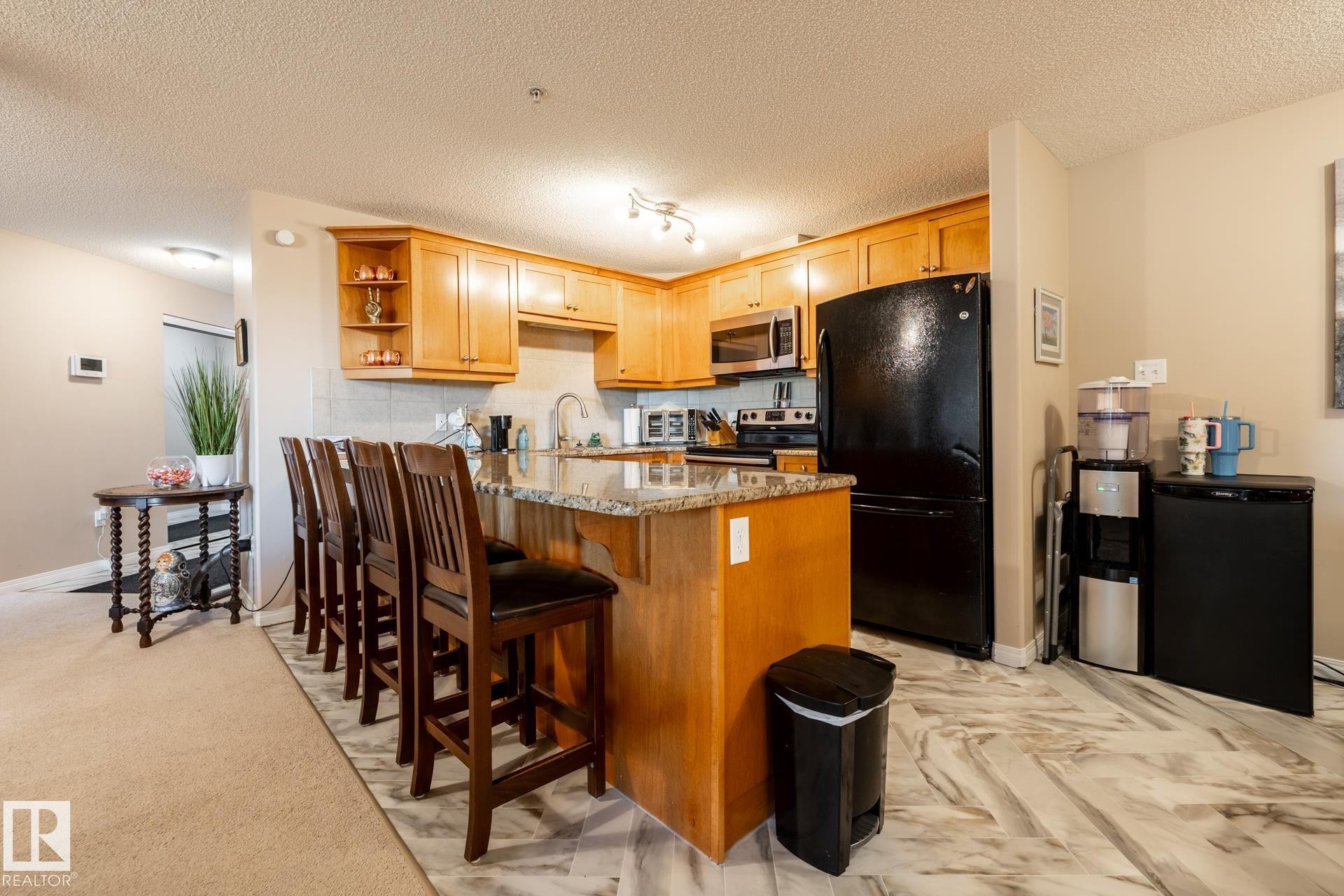 Kitchen featuring wood cabinetry, granite-finish countertops, black appliances, and a marble-look tile floor - 330 1520 Hammond Gate Gate, Edmonton, AB