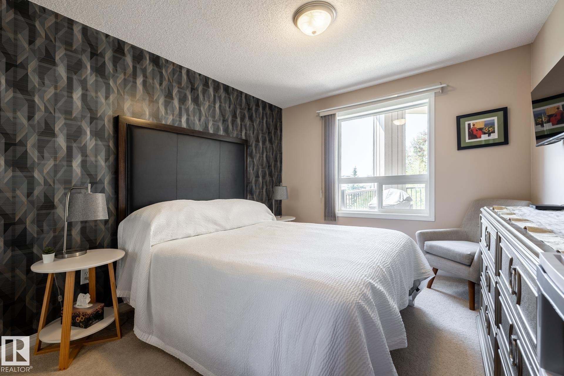 Bedroom featuring a geometric accent wall, large window, soft neutral wall paint, and a textured carpeted floor - 330 1520 Hammond Gate Gate, Edmonton, AB