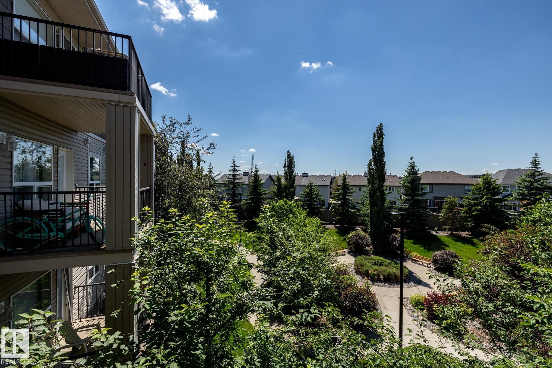 Private balcony with black railing overlooking a landscaped parkway featuring mature trees, paved pathways, and green spaces - 330 1520 Hammond Gate Gate, Edmonton, AB