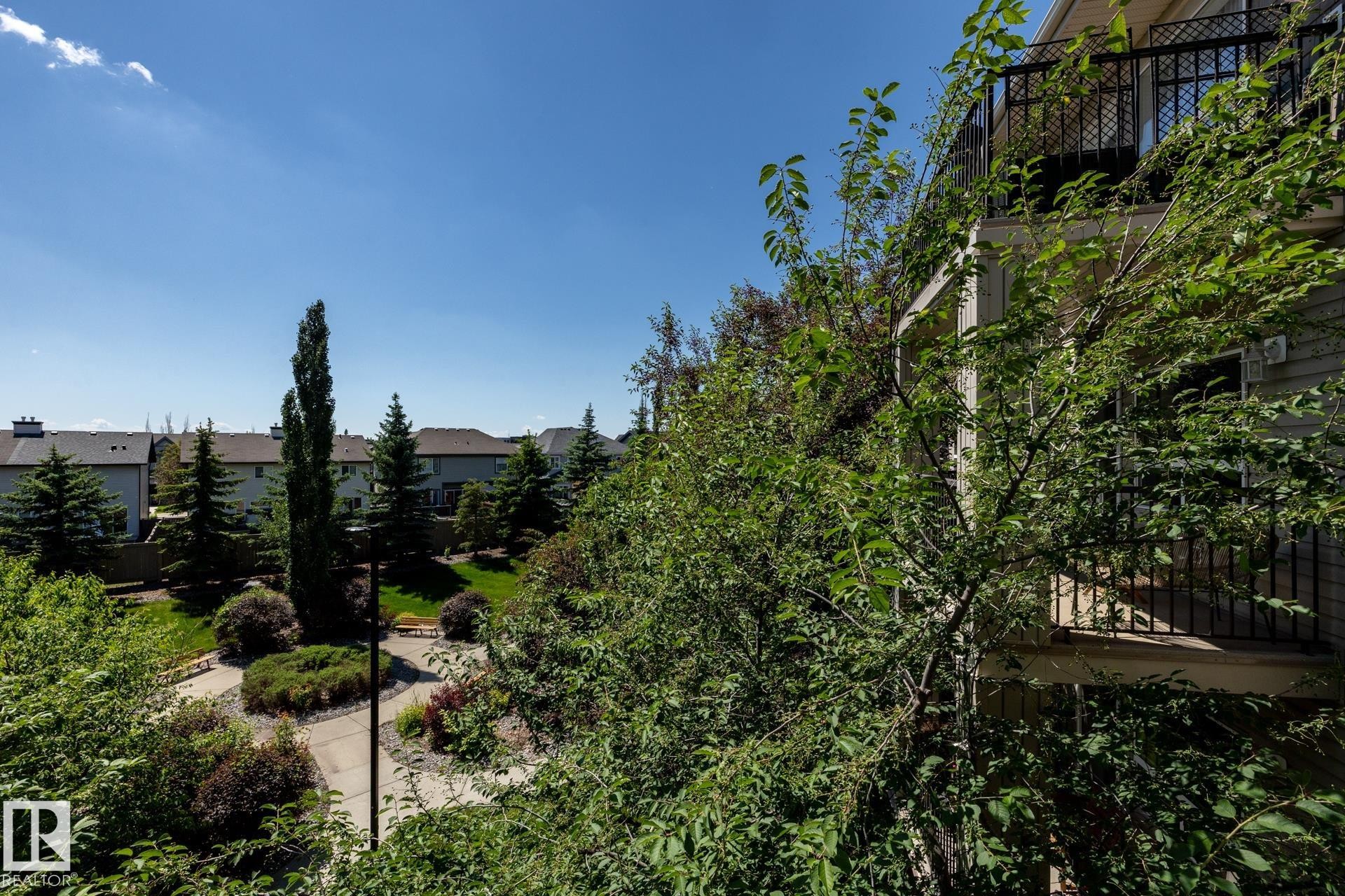 Private balcony with black wrought iron railing overlooking a landscaped green space with mature evergreen trees, paved walkways, and residential rooftops in the distance - 330 1520 Hammond Gate Gate, Edmonton, AB