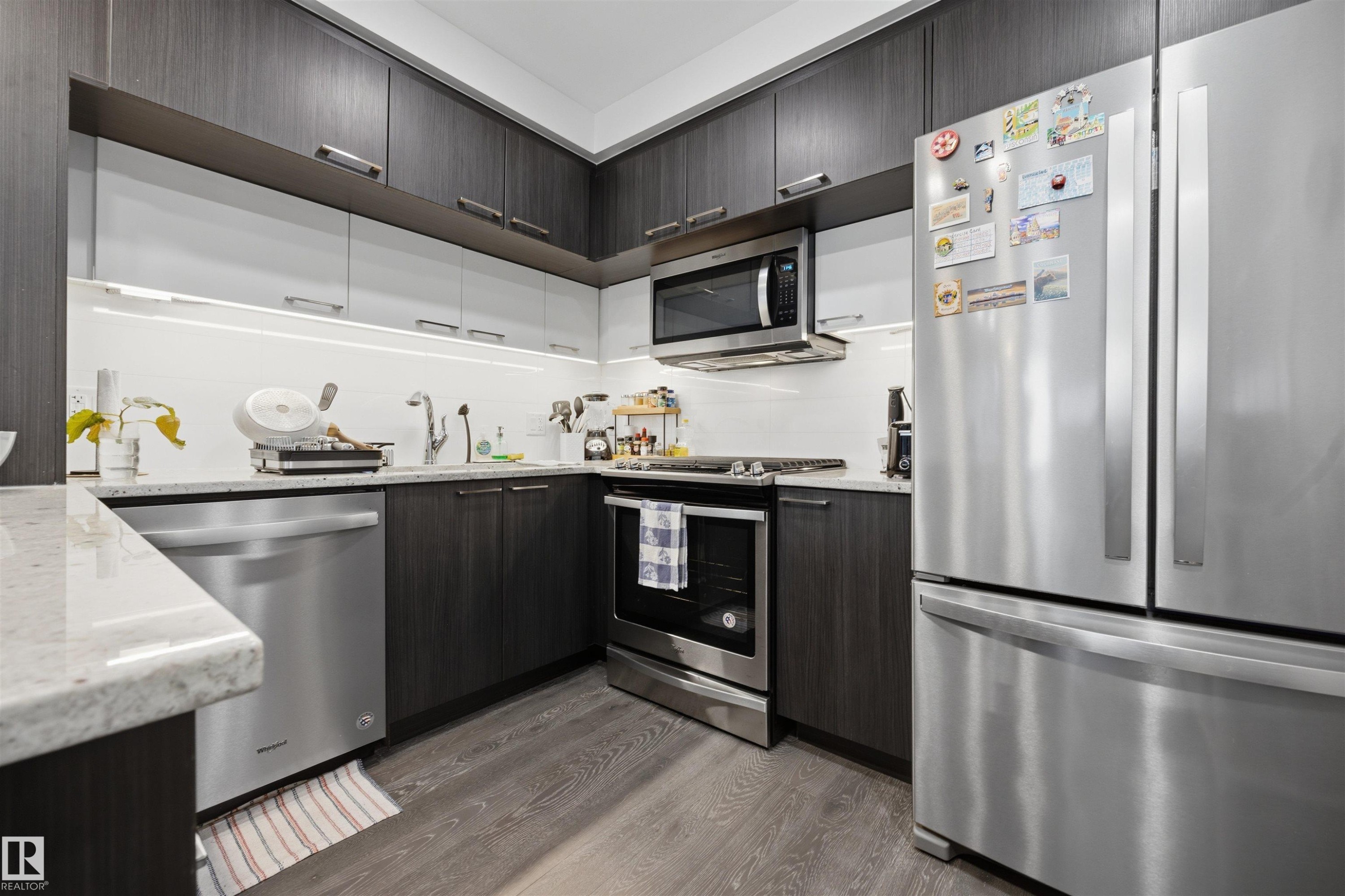 Modern kitchen featuring two-tone cabinetry, light-colored countertops, stainless steel appliances including a refrigerator, dishwasher, and microwave, and wood-finish flooring - #802 9720 106 St Nw, Edmonton, AB - Indoor Photo Showing Kitchen With Stainless Steel Kitchen With Upgraded Kitchen