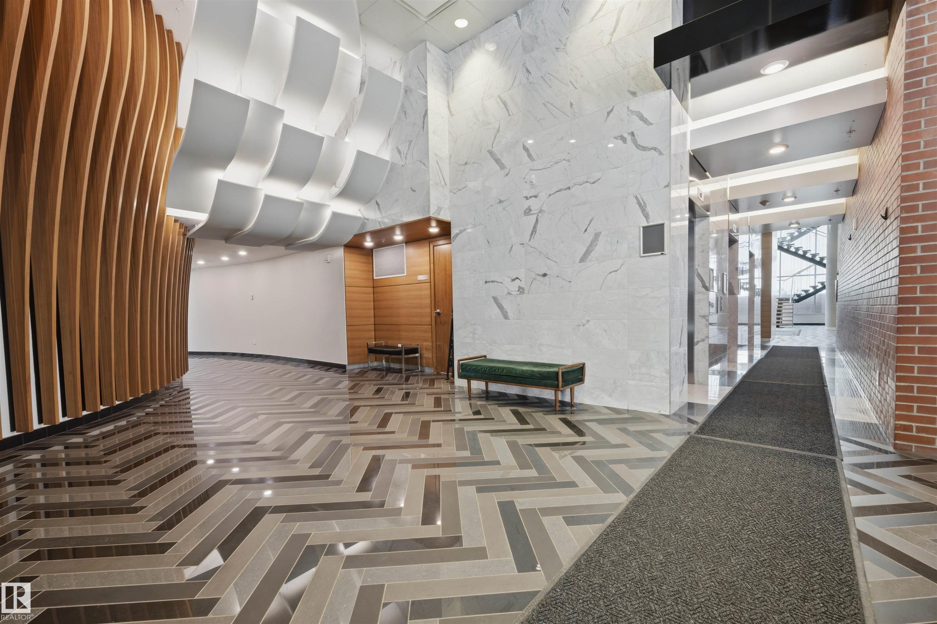 Lobby featuring a herringbone pattern floor, curved wood-panel wall, recessed lighting, and a white marble-look accent wall - #802 9720 106 St Nw, Edmonton, AB - Indoor Photo Showing Other Room