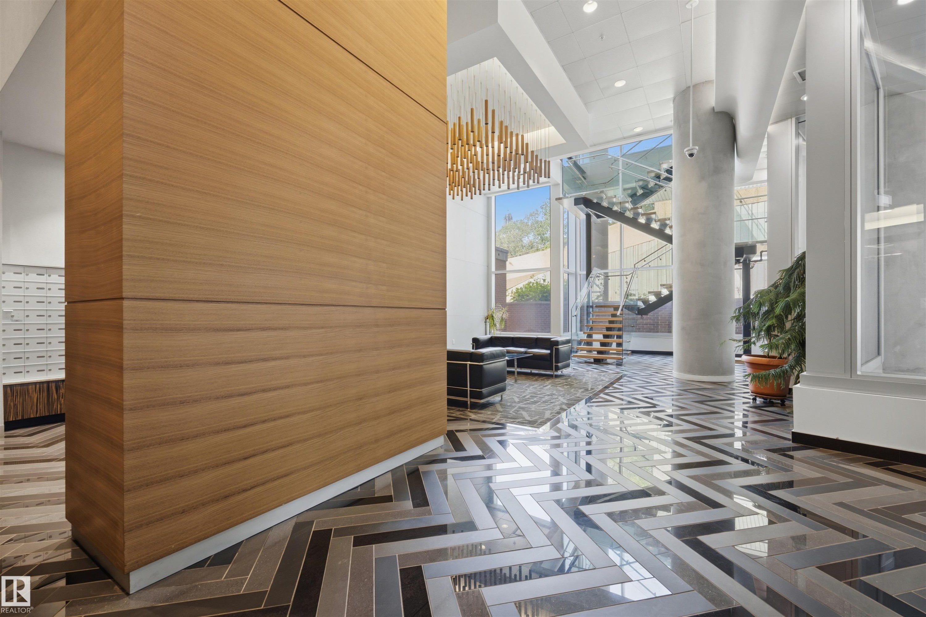 Lobby entrance featuring chevron-patterned flooring, a tall wood-paneled wall, a multi-tiered chandelier, and a prominent glass and metal staircase - #802 9720 106 St Nw, Edmonton, AB - Indoor Photo Showing Other Room