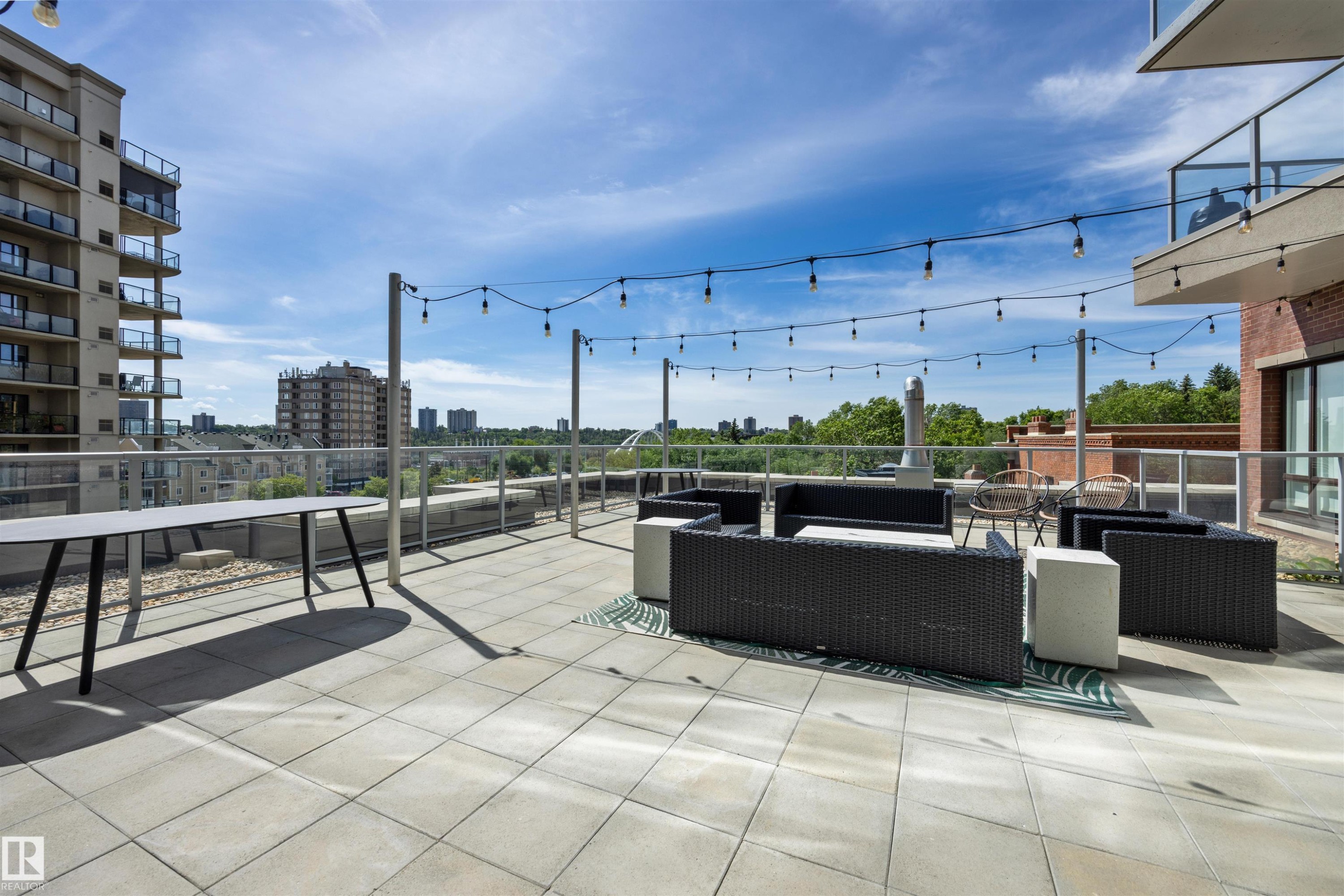 Expansive rooftop terrace with large format paver flooring, string lighting, glass and metal railings, and brick architectural elements - #802 9720 106 St Nw, Edmonton, AB - Outdoor With Balcony
