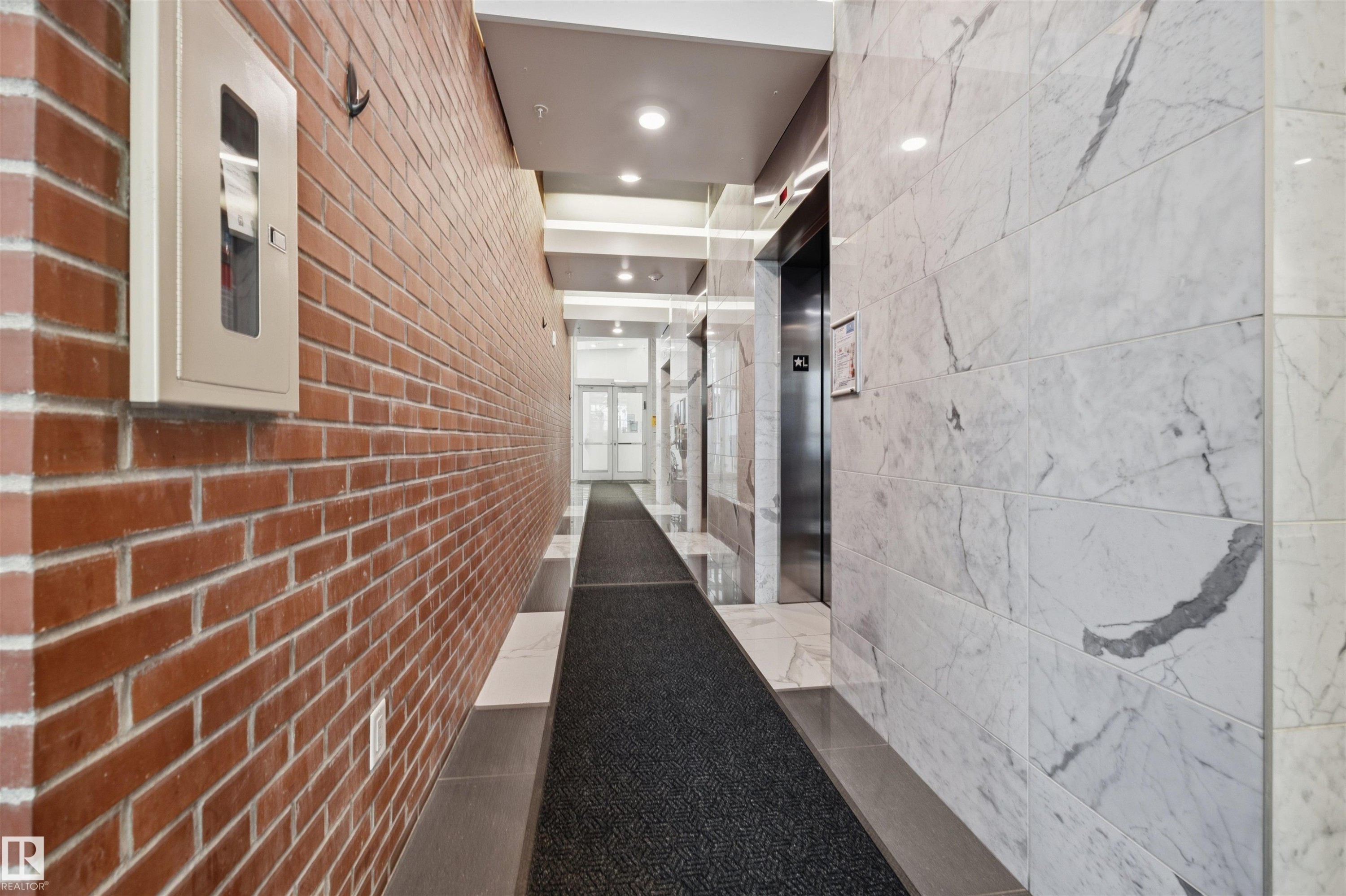 Building hallway featuring a brick accent wall, marble-patterned wall panels, recessed lighting, and elevator access - #802 9720 106 St Nw, Edmonton, AB - Indoor
