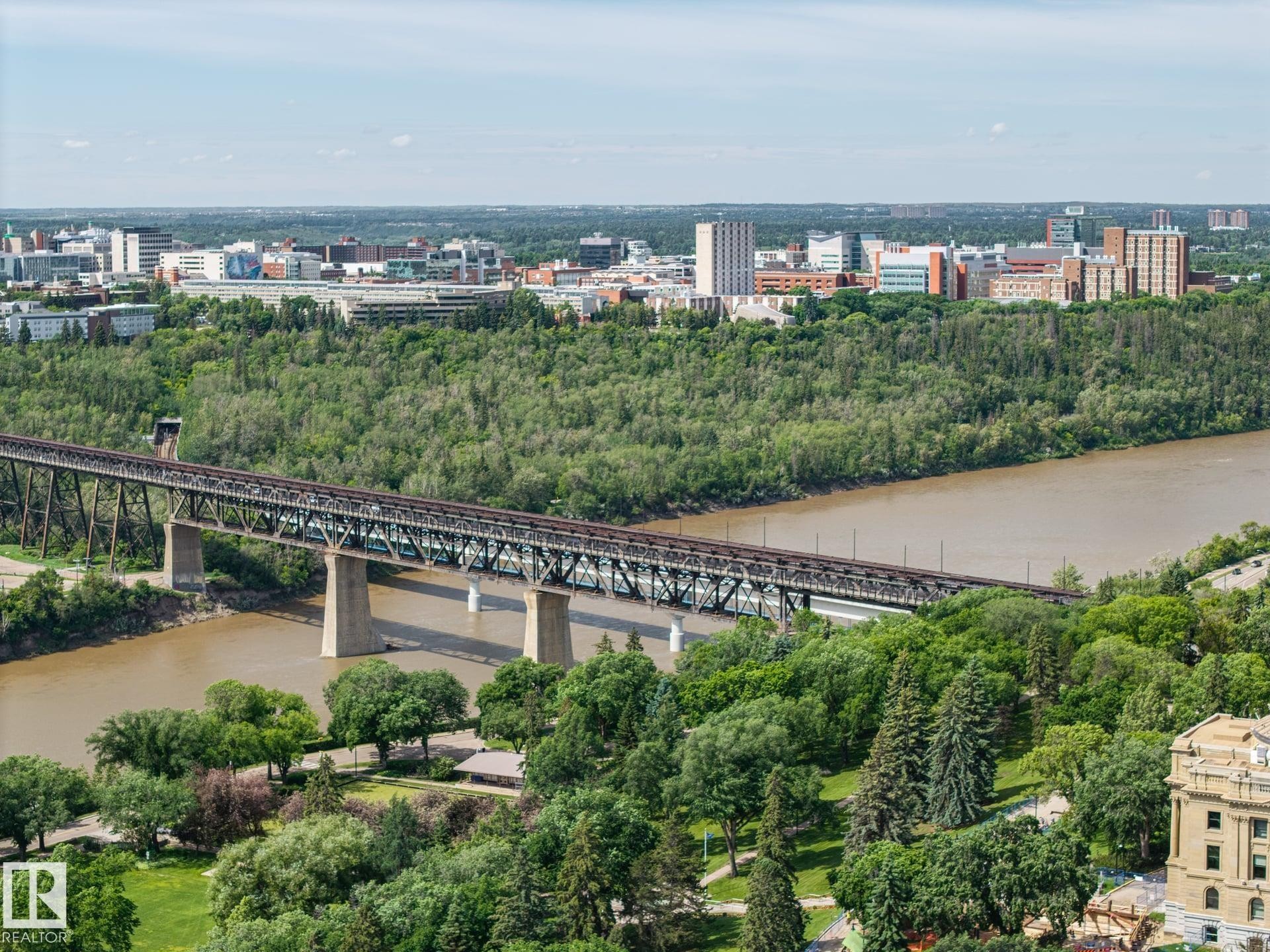 Expansive urban landscape featuring a wide river, a prominent railway bridge, abundant mature trees, and a distant city skyline with multiple buildings - #802 9720 106 St Nw, Edmonton, AB - Outdoor With View