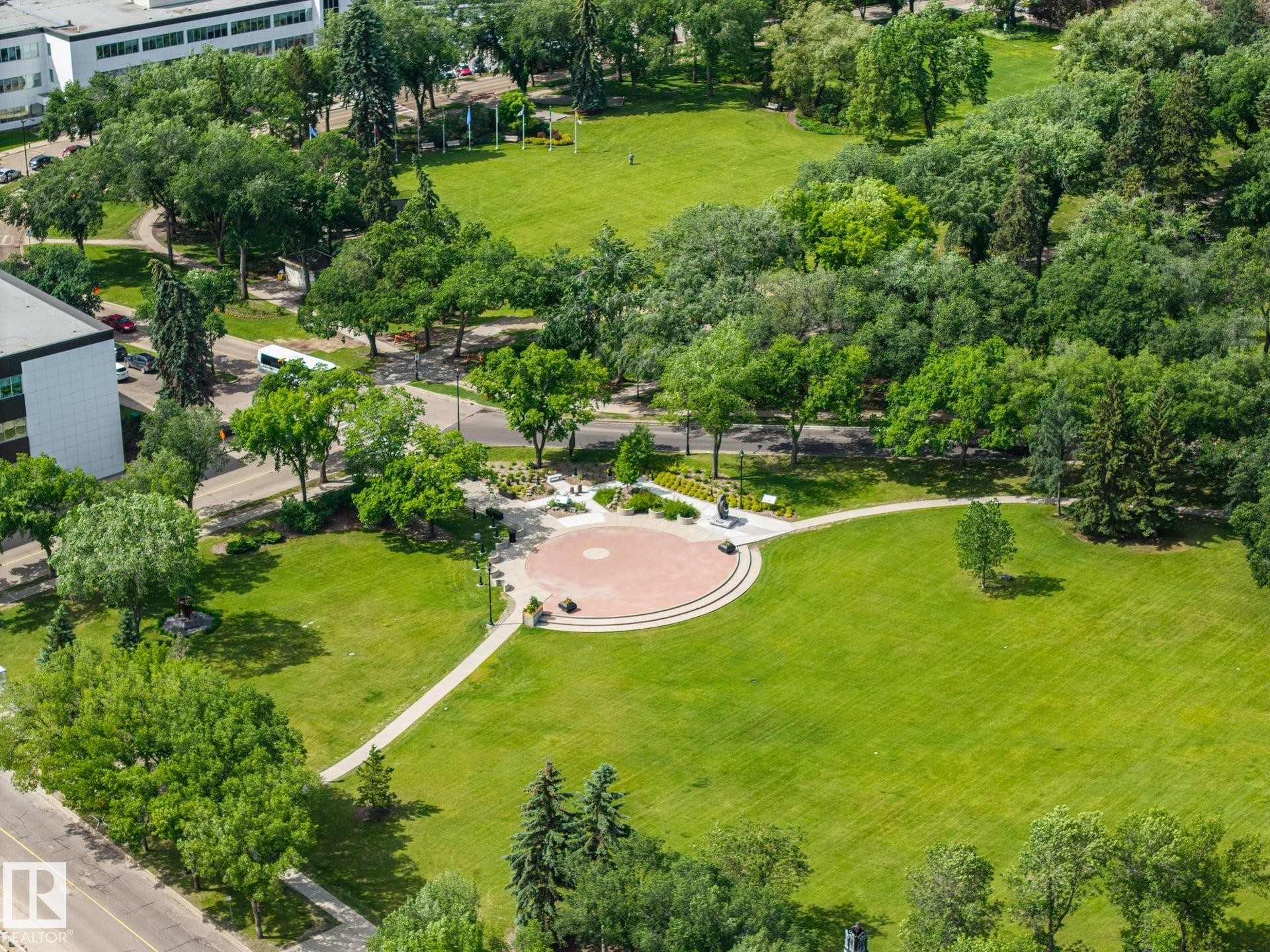 Expansive park grounds featuring lush green lawns, mature trees, paved walking paths, and a circular brick patio with tiered seating - #802 9720 106 St Nw, Edmonton, AB - Outdoor