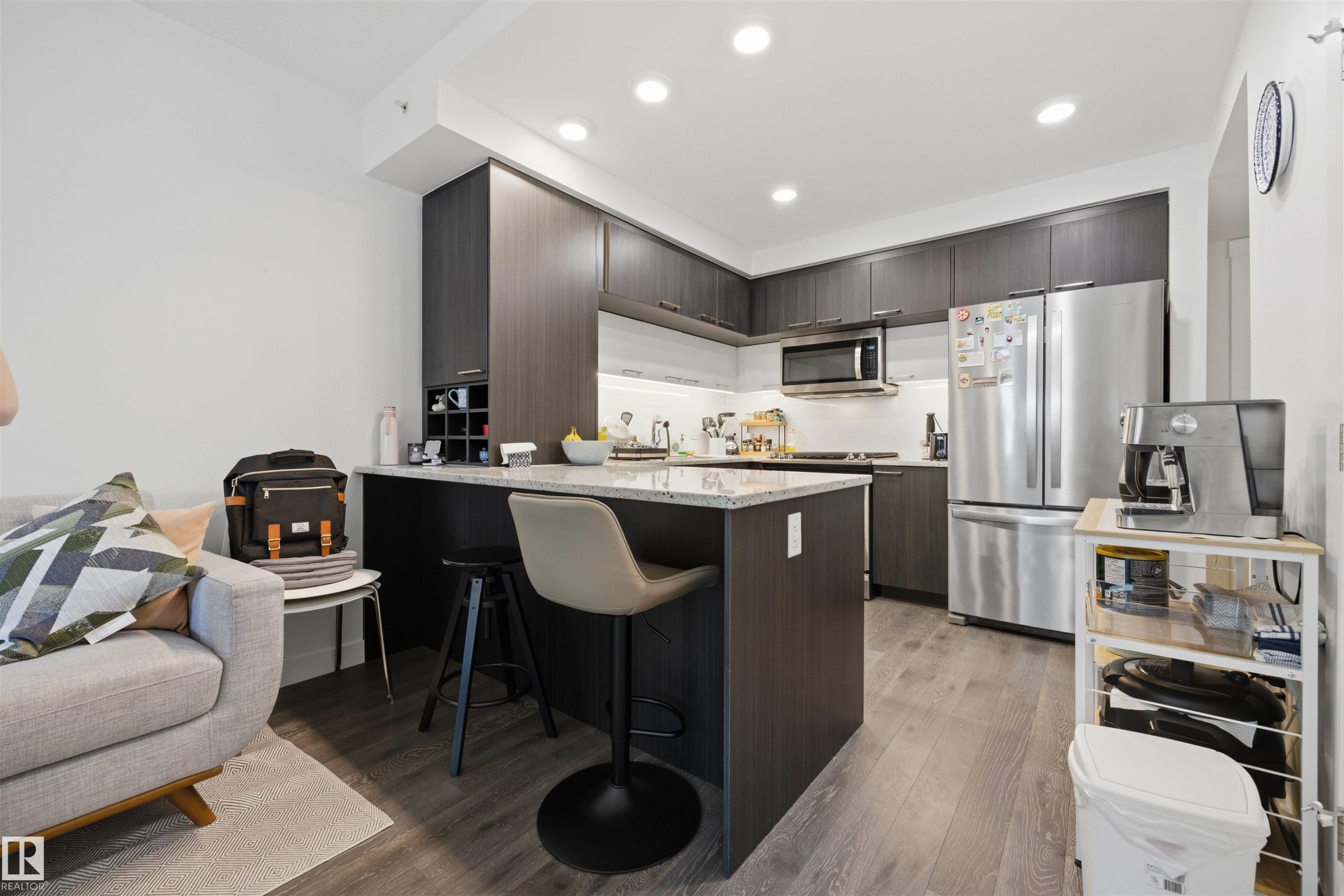 Contemporary kitchen featuring dark wood-finish cabinetry, a stone countertop island with seating, stainless steel appliances, and recessed lighting - #802 9720 106 St Nw, Edmonton, AB - Indoor Photo Showing Kitchen With Stainless Steel Kitchen With Upgraded Kitchen