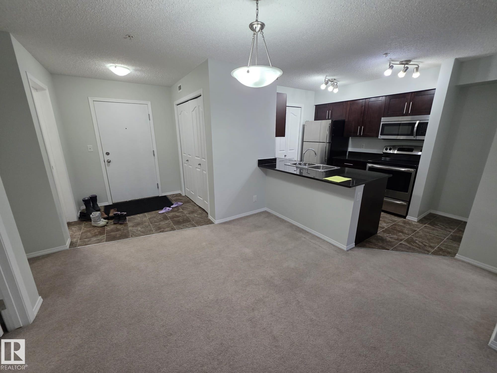 Open-concept living space featuring a kitchen with dark wood-finish cabinetry, stainless steel appliances, and a breakfast bar with a dark countertop - 206 12045 22 Avenue, Edmonton, AB