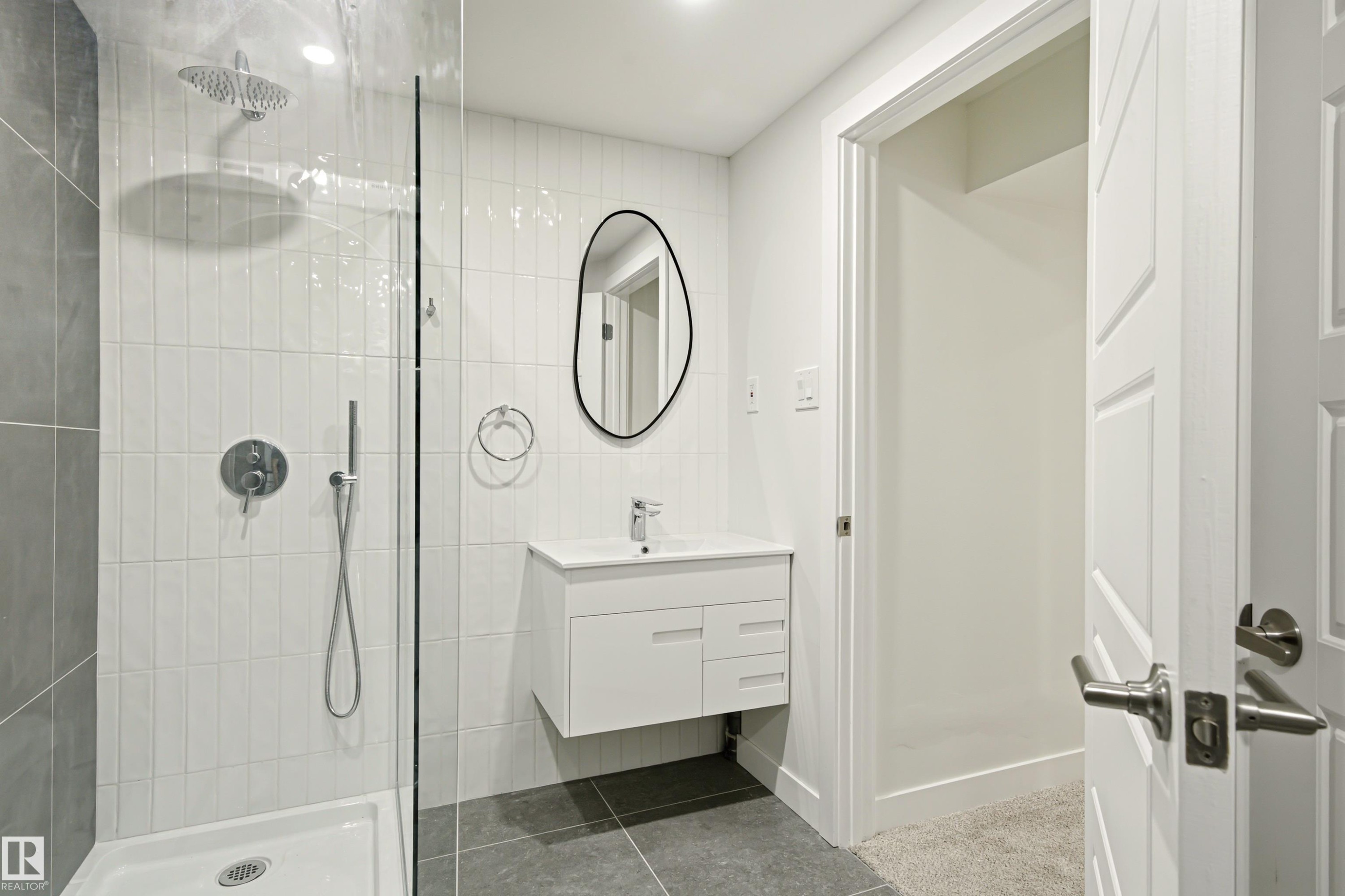 Bathroom featuring a glass-enclosed shower with a rainfall shower head and handheld sprayer, white vertical subway tile, a floating vanity with a white countertop and integrated sink, and an organic-shaped black-framed mirror - 18217 84 Avenue Nw, Edmonton, AB