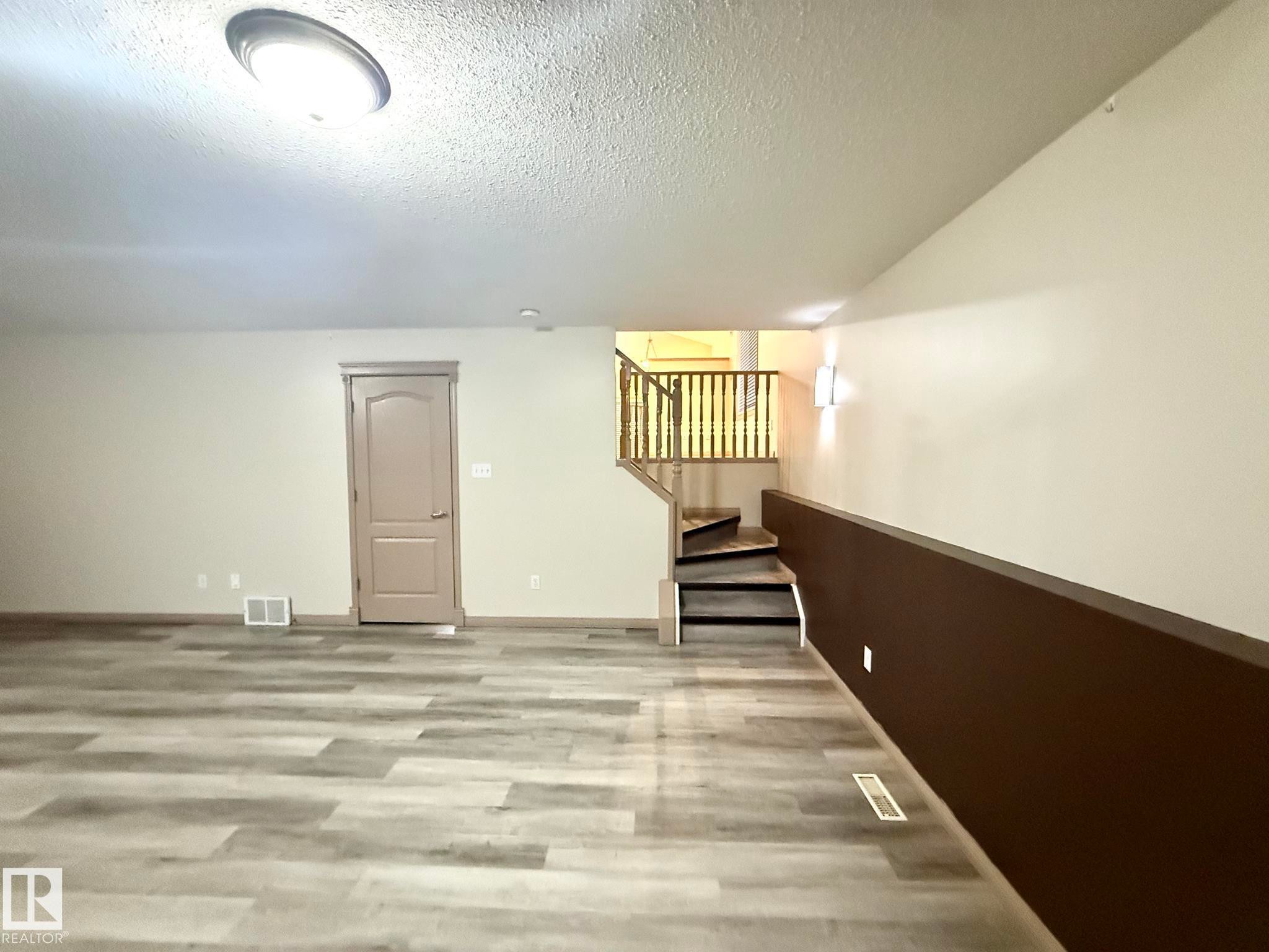 Spacious lower-level room featuring wood-finish flooring, neutral wall tones, a paneled interior door, and a staircase with wood railings and treads - 1978 Tomlinson Green Green, Edmonton, AB