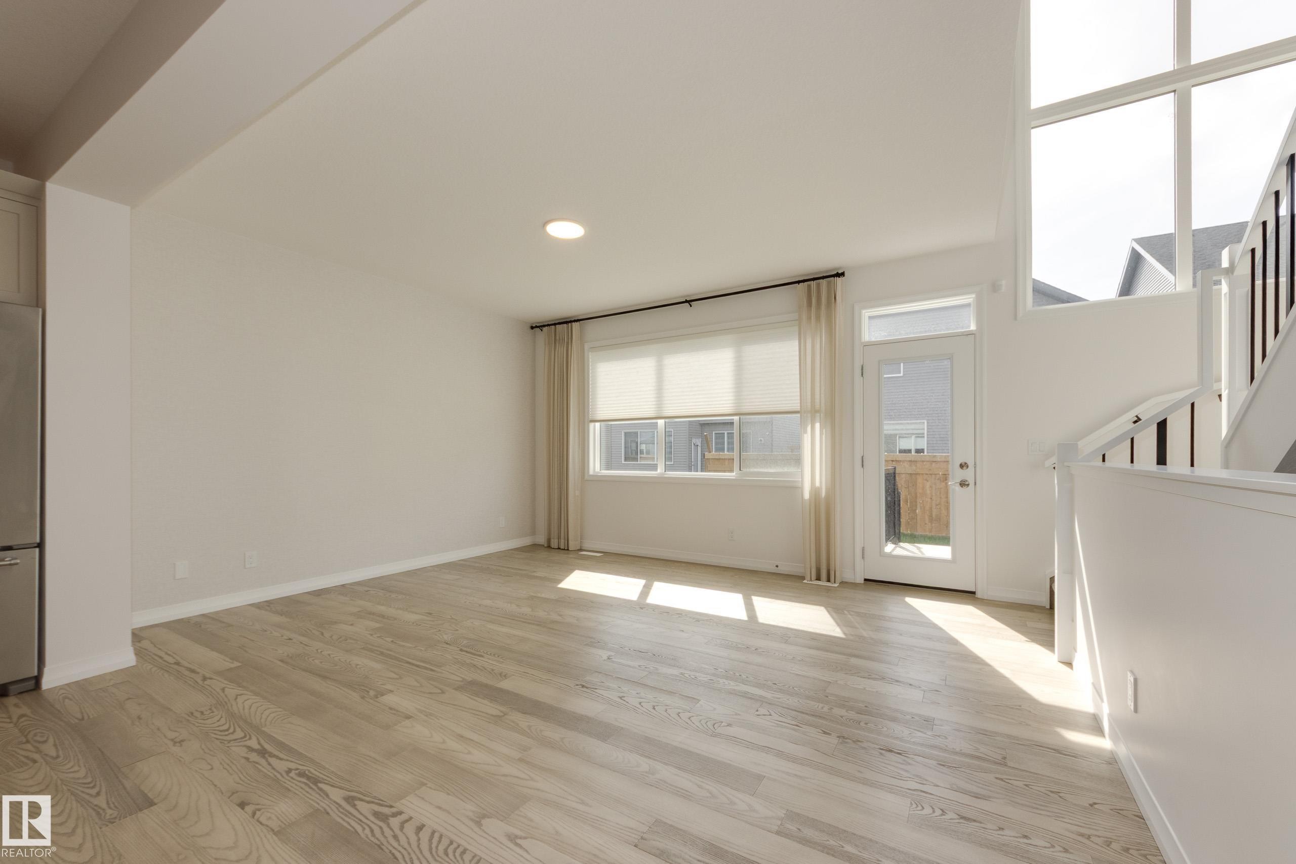 Spacious interior featuring light wood-finish flooring, a large window with roller shades, and a glass-paneled exterior door - 3249 Dixon Way Sw, Edmonton, AB