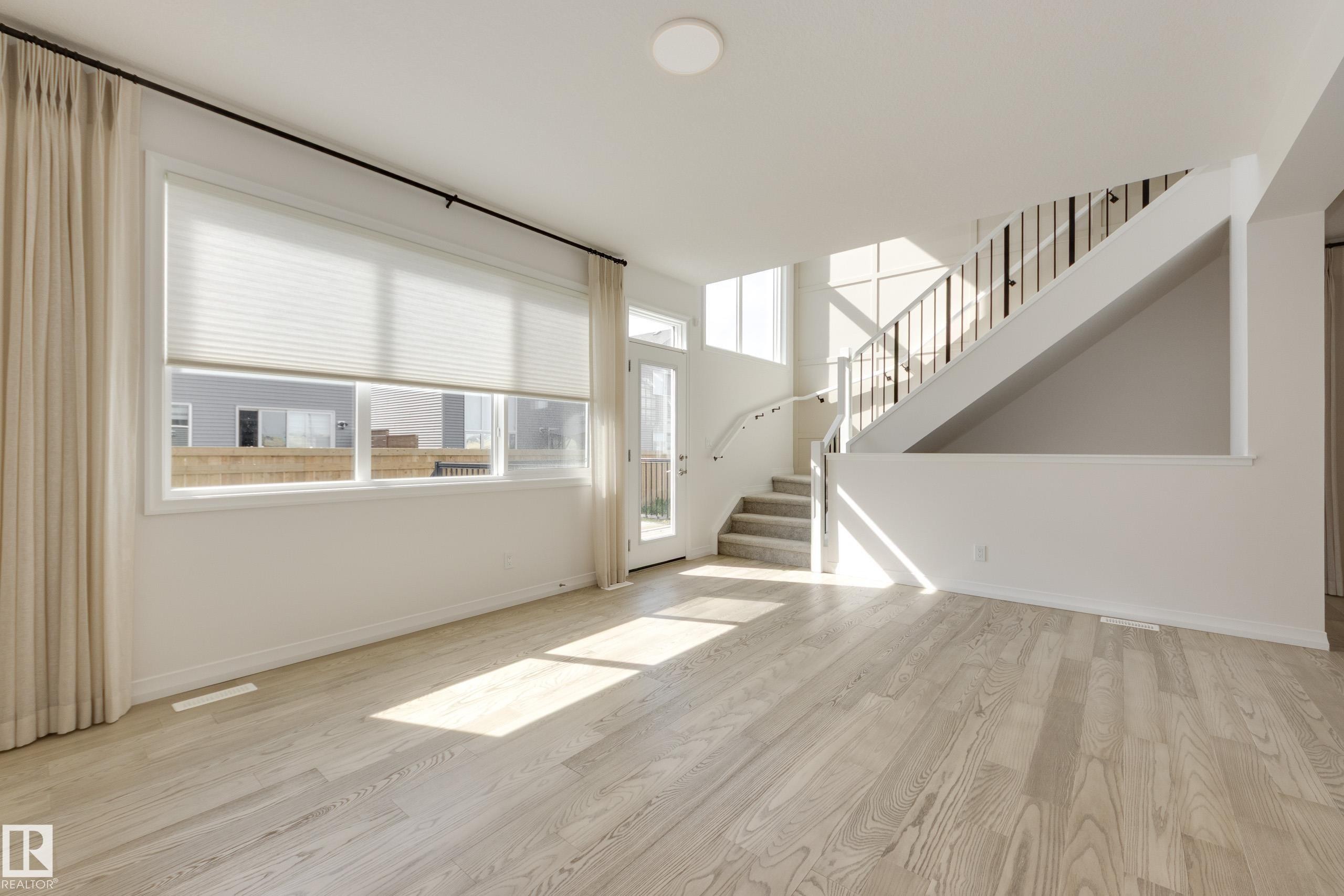 Open concept living space featuring light wood-finish flooring, a contemporary staircase with metal spindles, and a multi-pane glass door - 3249 Dixon Way Sw, Edmonton, AB