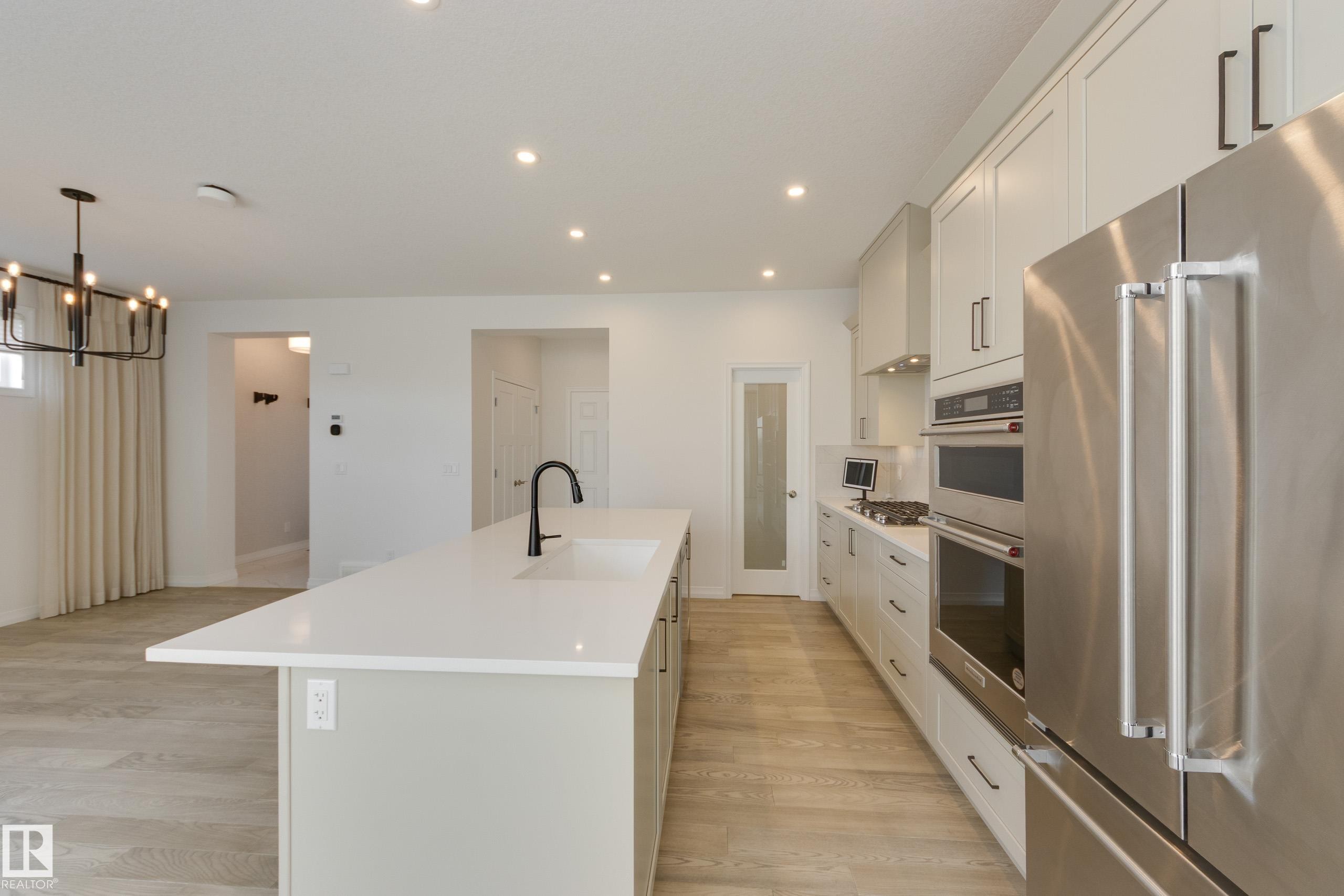 Modern kitchen featuring a large island with an undermount sink and black faucet, light wood-finish flooring, recessed lighting, white shaker cabinetry with dark hardware, and stainless steel appliances including a built-in oven and refrigerator - 3249 Dixon Way Sw, Edmonton, AB