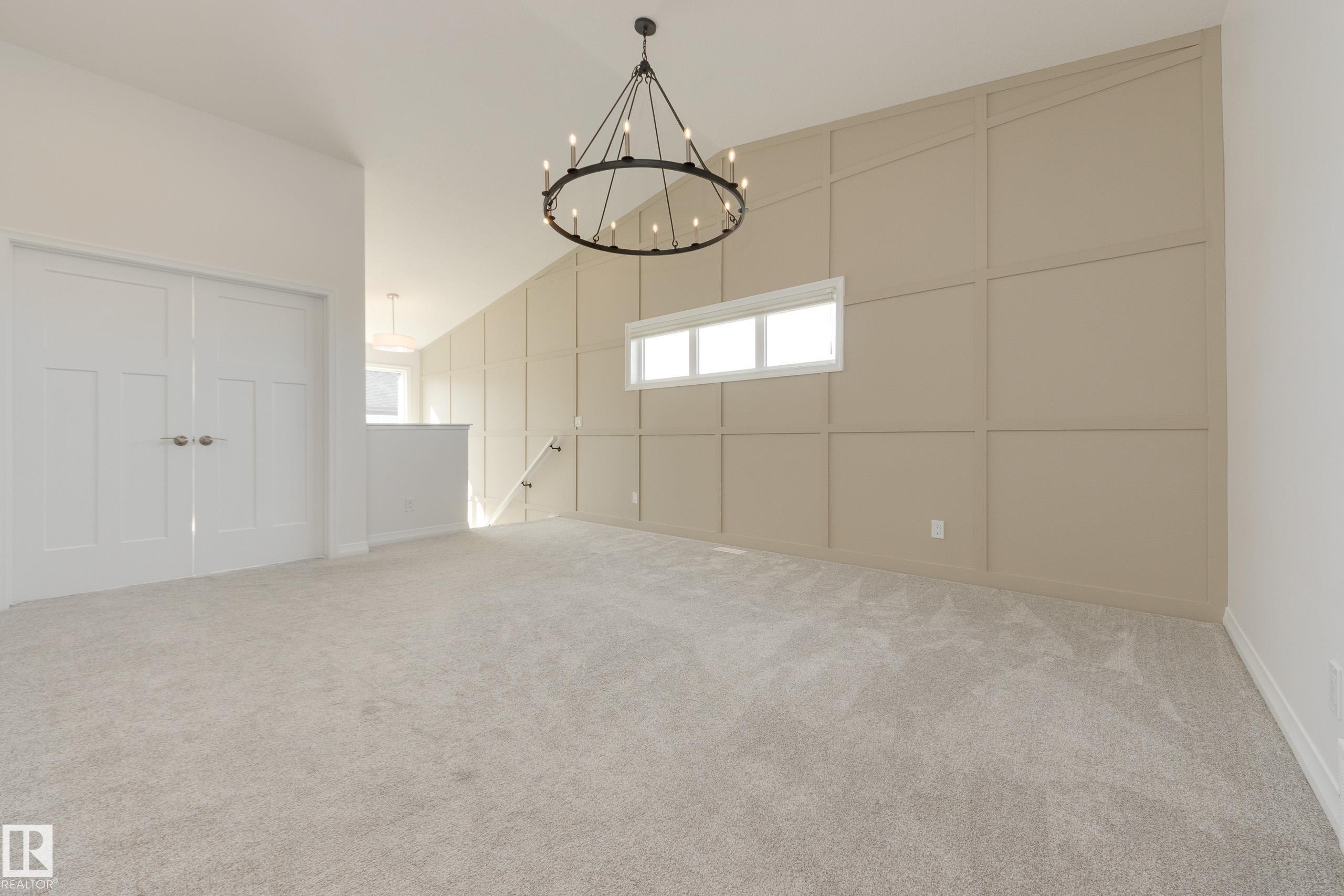 Spacious room featuring a decorative accent wall, a multi-pane horizontal window, and a large chandelier - 3249 Dixon Way Sw, Edmonton, AB