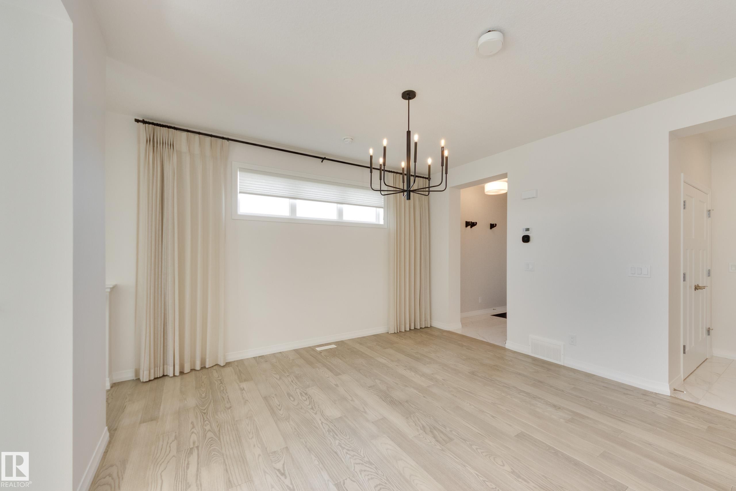 Spacious room featuring light wood-finish flooring, a modern black chandelier, and a large window with roller shades - 3249 Dixon Way Sw, Edmonton, AB