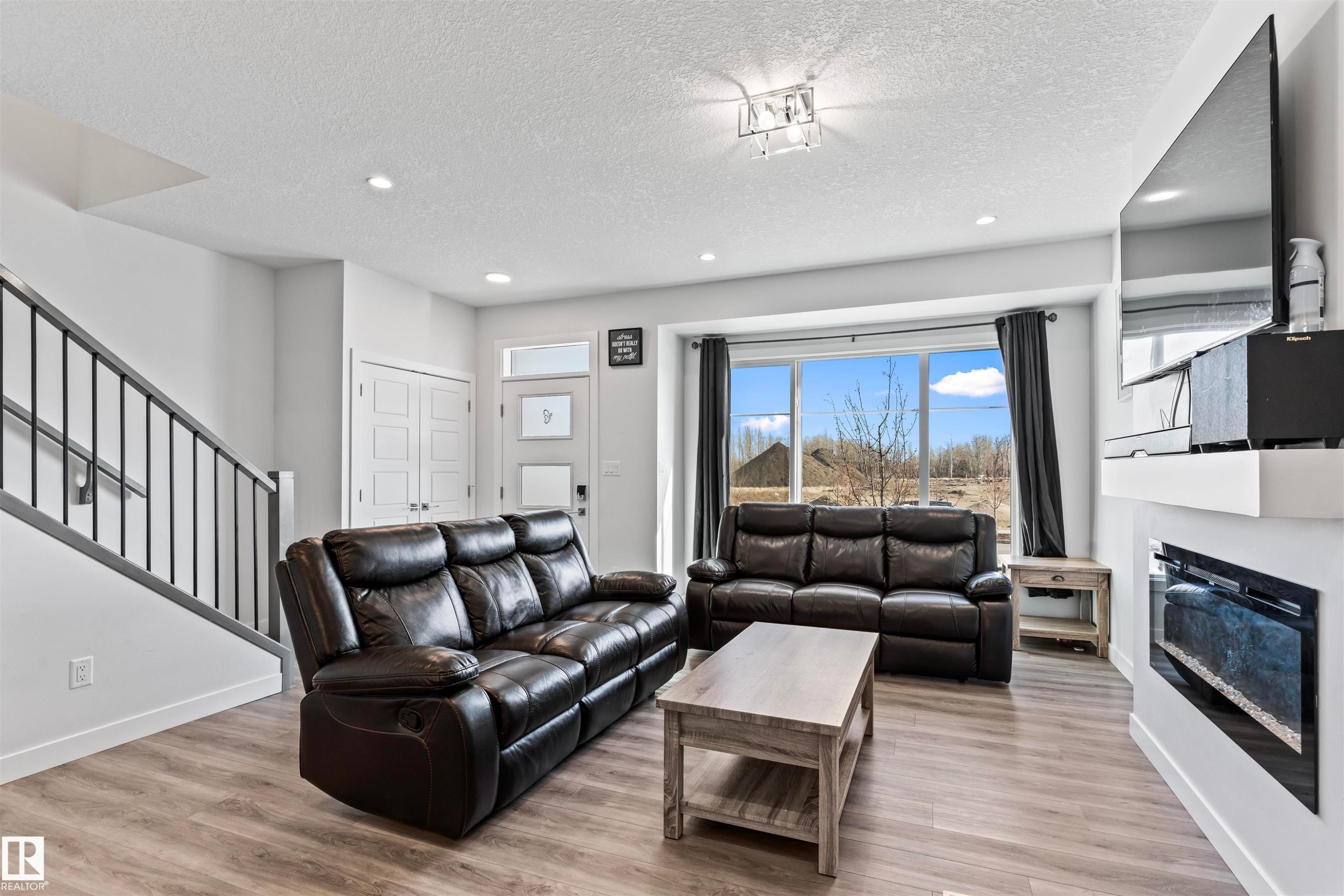 Bright living area featuring wood-finish flooring, a contemporary electric fireplace with a white mantel, and a prominent bay window - 8046 Kiriak Link, Edmonton, AB - Indoor Photo Showing Living Room With Fireplace