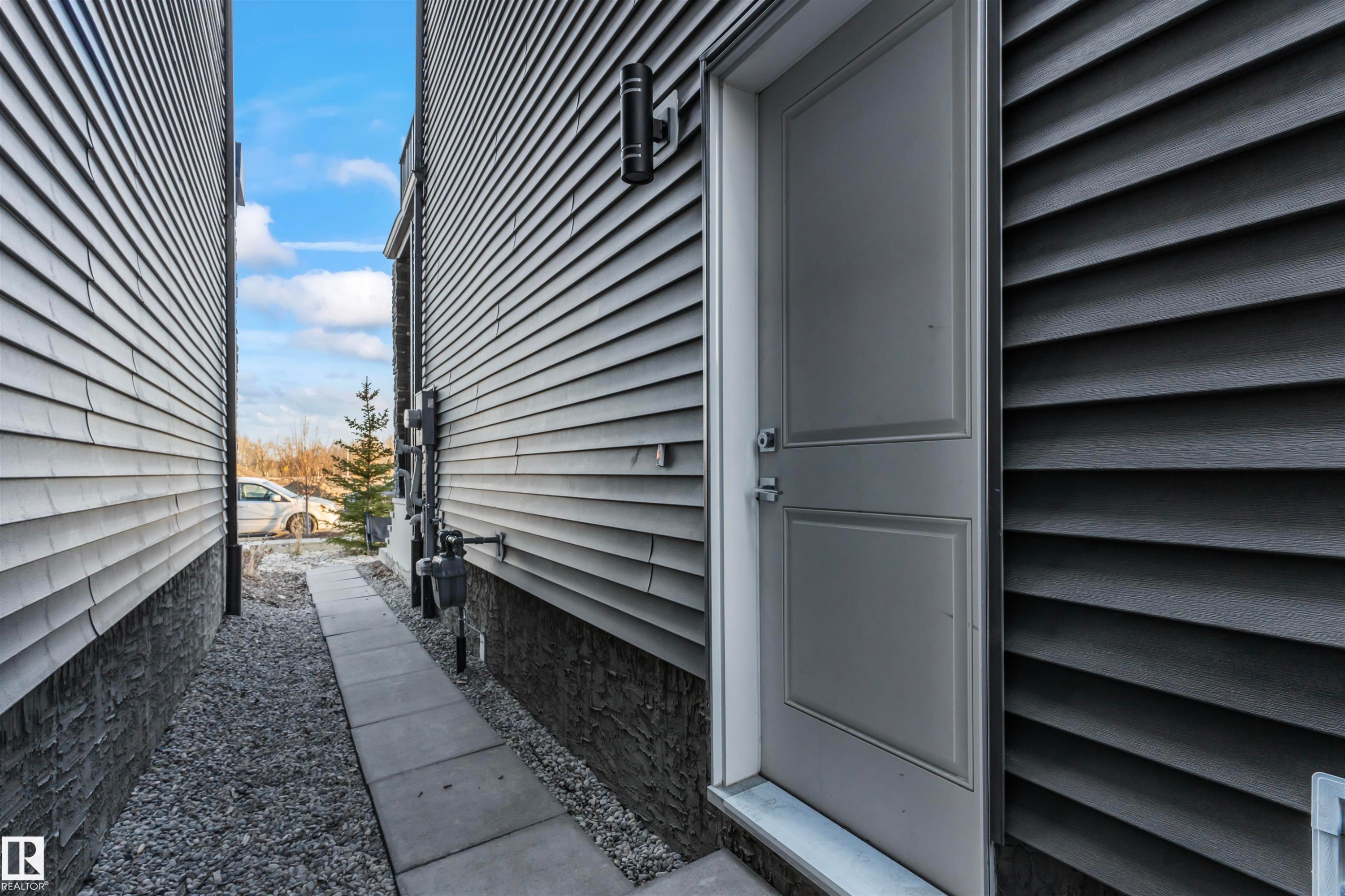 Exterior entry featuring a light gray door with panel detailing, dark gray horizontal siding, and a matte black wall-mounted light fixture - 8046 Kiriak Link, Edmonton, AB - Outdoor With Exterior