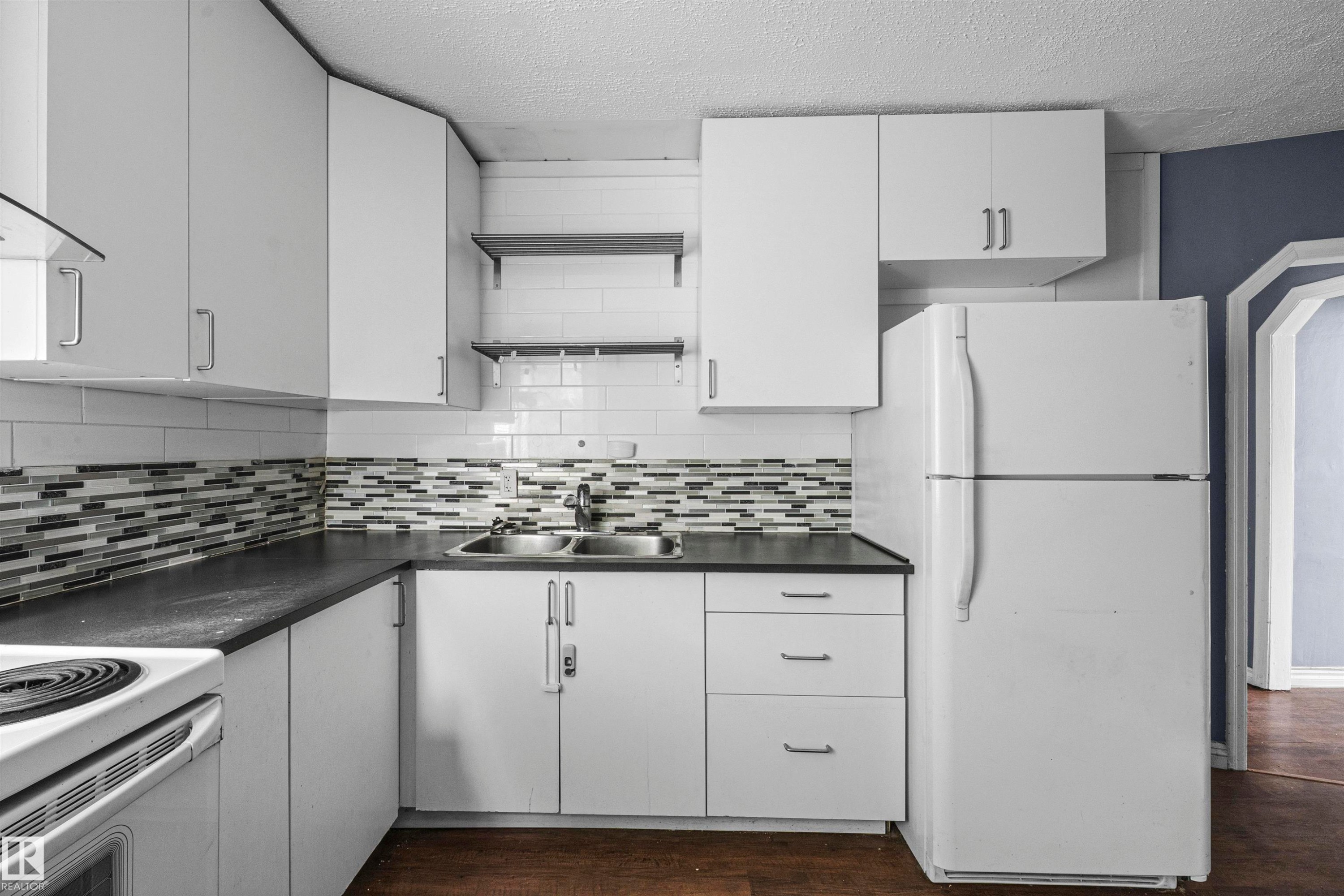 Kitchen featuring white cabinetry with silver-tone hardware, a dual basin stainless steel sink, dark countertops, and a subway tile backsplash with an accent mosaic - 7520 106 Street, Edmonton, AB