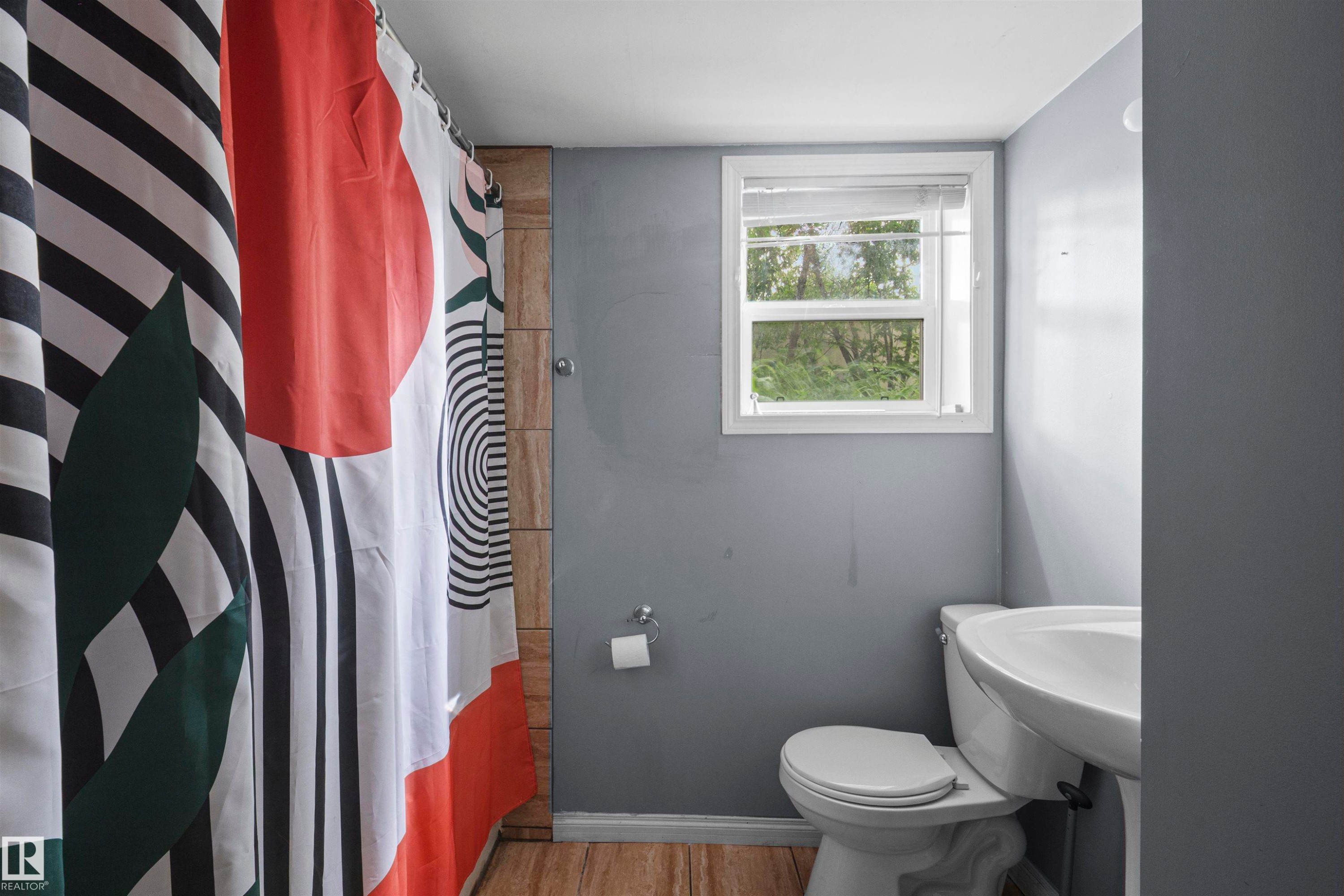 Bathroom featuring a pedestal sink, toilet, single window, wood-finish flooring, and light gray wall paint - 7520 106 Street, Edmonton, AB