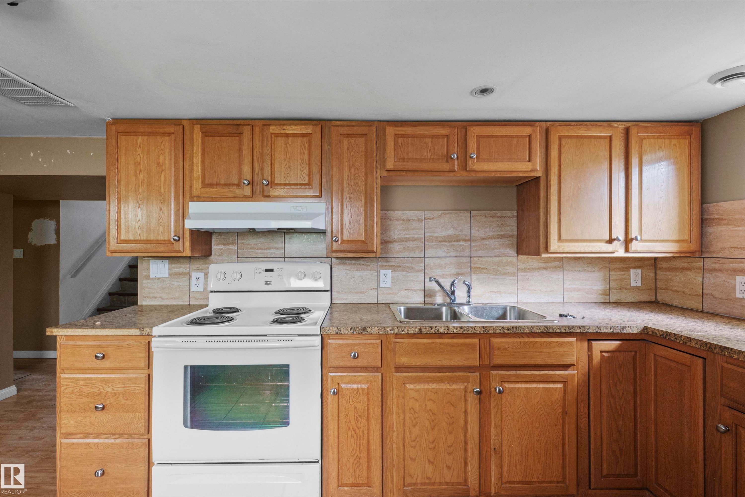 Kitchen featuring wood cabinetry, stone-finish countertops, ceramic tile backsplash, a double basin stainless steel sink, and a white electric range with a coordinating range hood - 7520 106 Street, Edmonton, AB