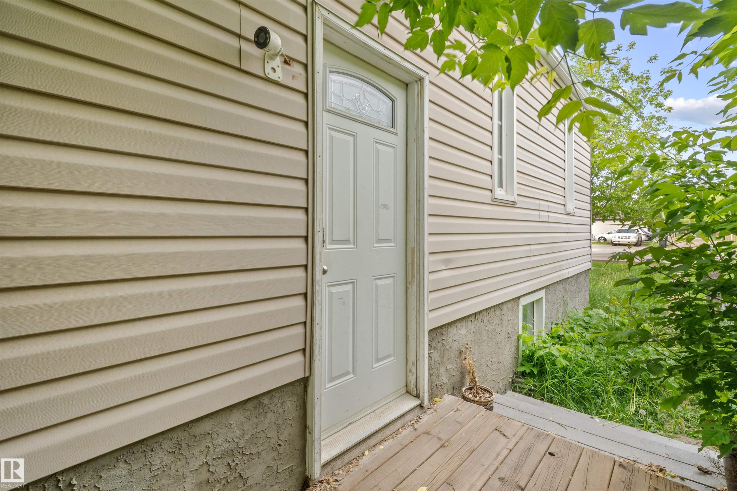 Tan horizontal siding exterior with a white paneled entry door featuring an arched window insert, set on a wooden deck - 7520 106 Street, Edmonton, AB