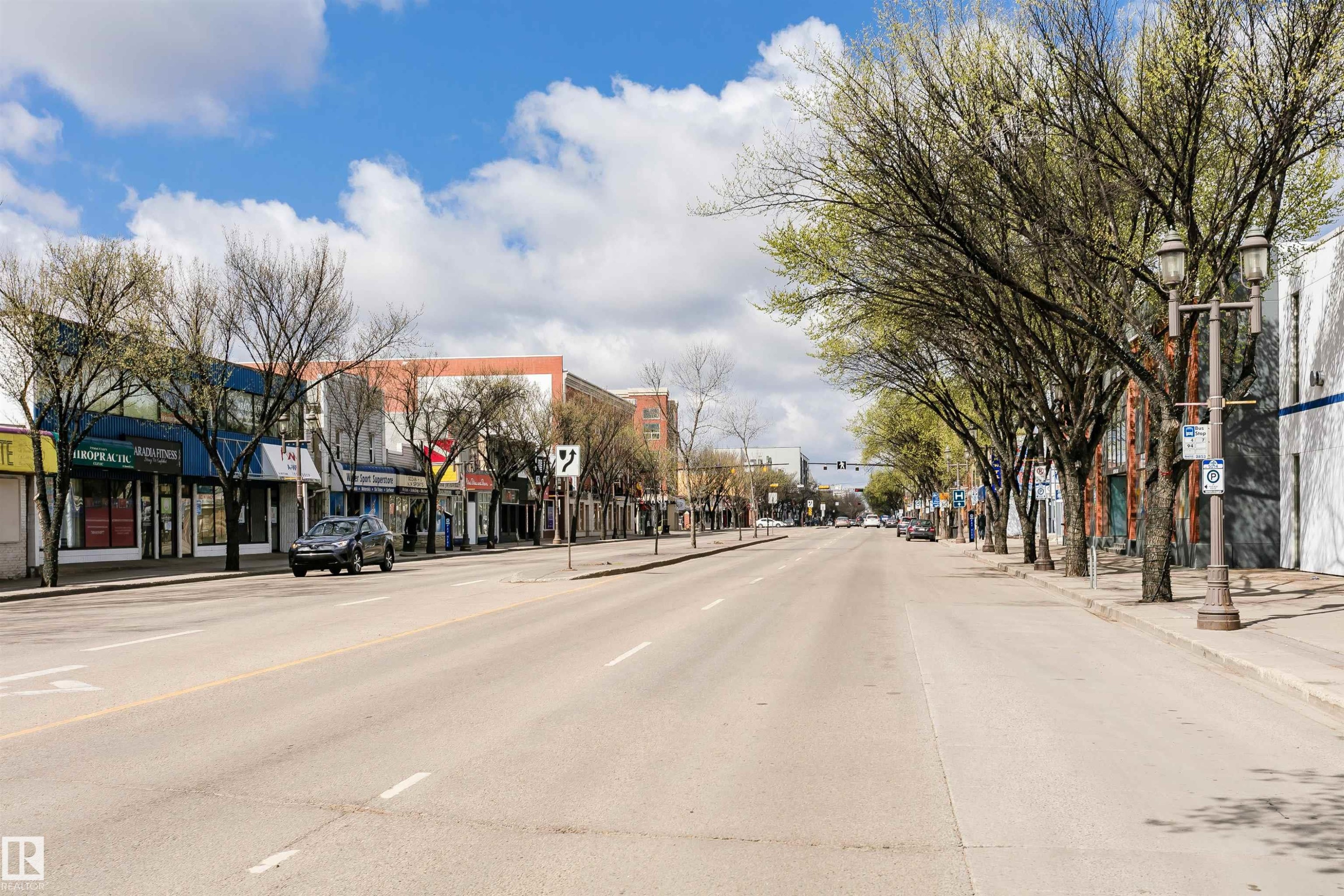 Commercial streetscape featuring multiple retail storefronts with large display windows, a wide paved road, and established trees lining the sidewalks - 10035 81 Avenue Nw, Edmonton, AB - Outdoor