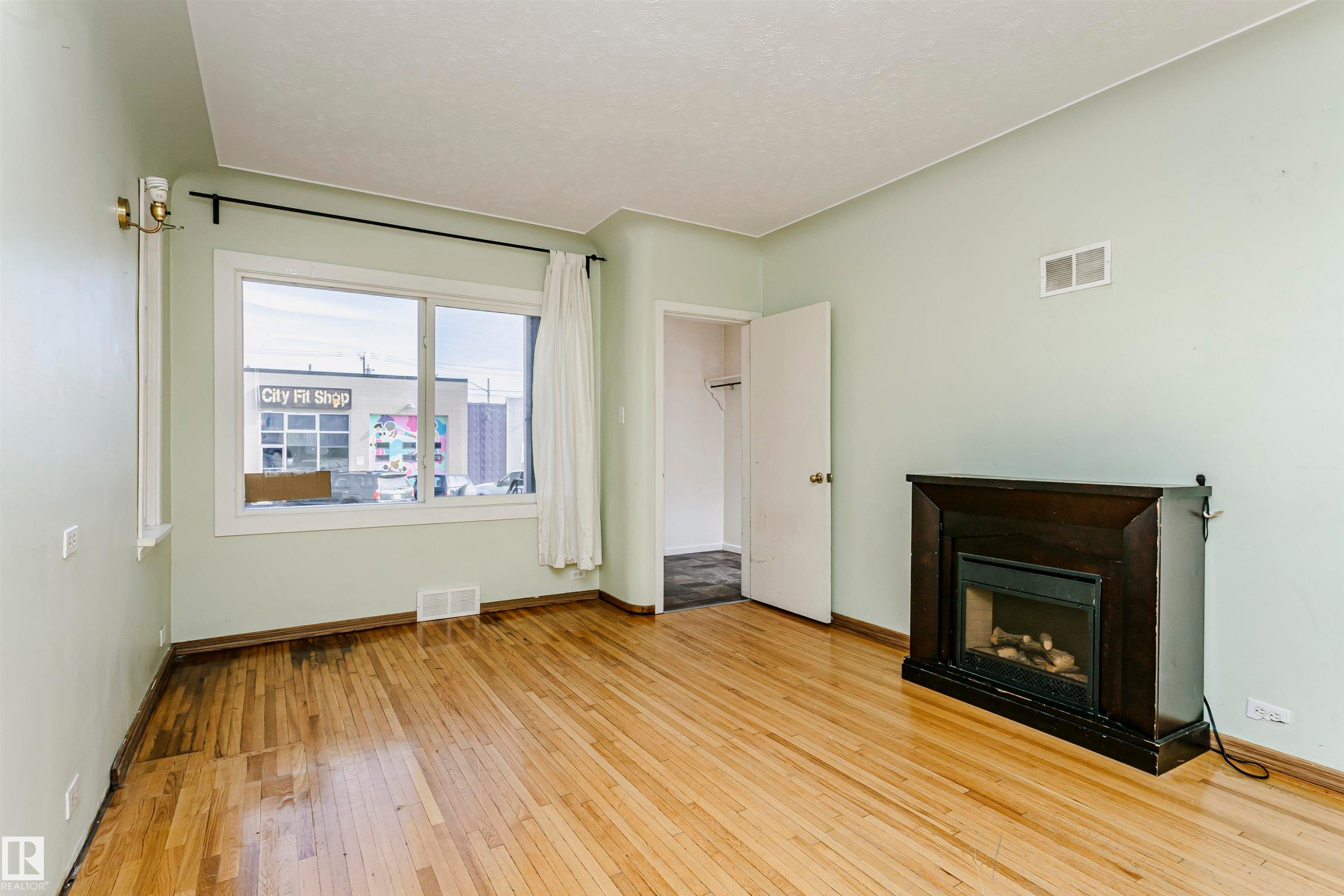 Hardwood flooring and a dark wood-finish fireplace frame this room, featuring light-colored walls and a window with white trim - 10035 81 Avenue Nw, Edmonton, AB - Indoor Photo Showing Other Room With Fireplace