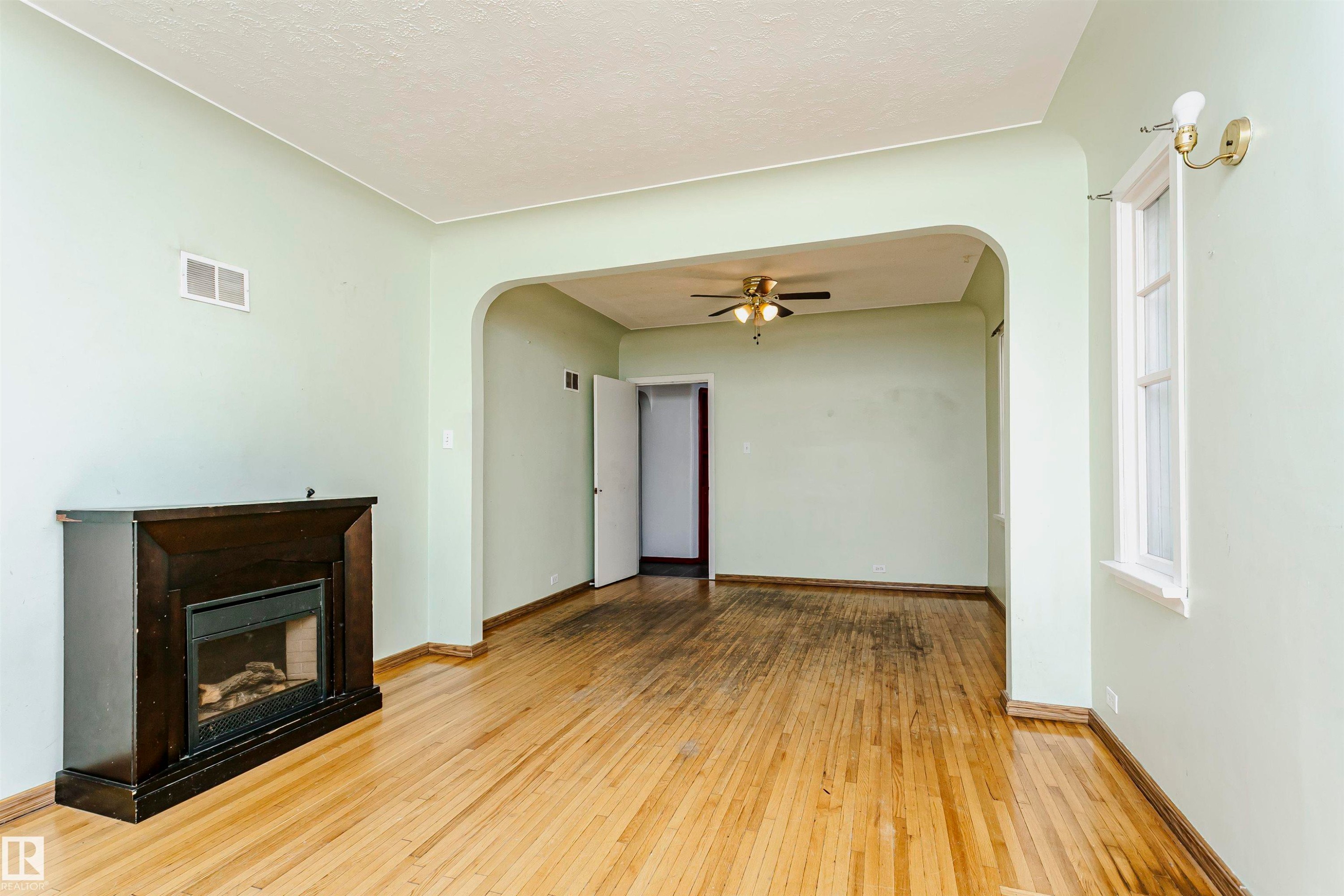Hardwood flooring extends throughout this space, featuring an arched doorway and a dark wood-finish mantel with an electric fireplace insert - 10035 81 Avenue Nw, Edmonton, AB - Indoor With Fireplace