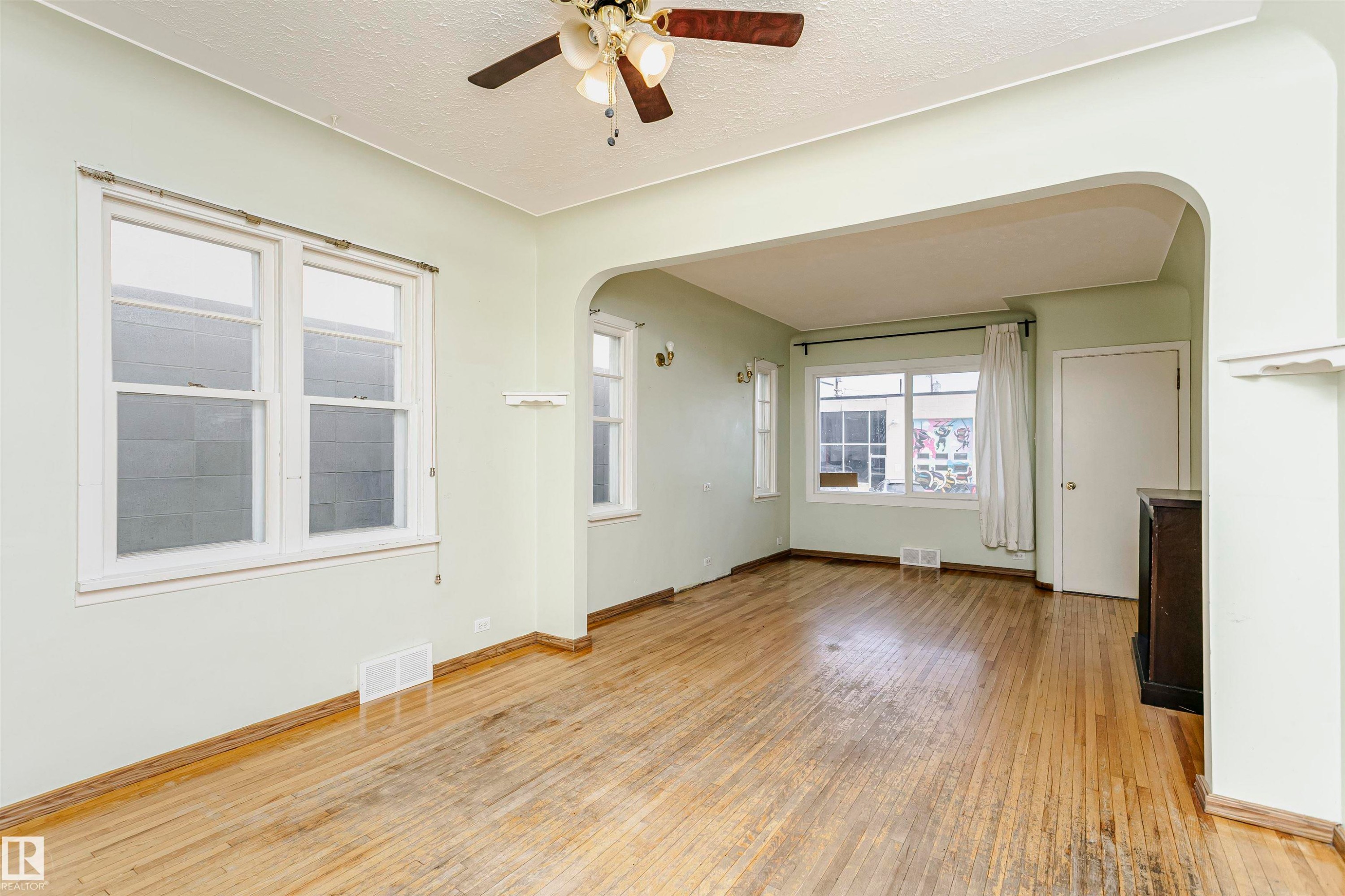 Open-concept living area featuring wood-finish flooring, multiple windows, a ceiling fan with integrated lighting, and an arched entryway to an additional room - 10035 81 Avenue Nw, Edmonton, AB - Indoor Photo Showing Other Room