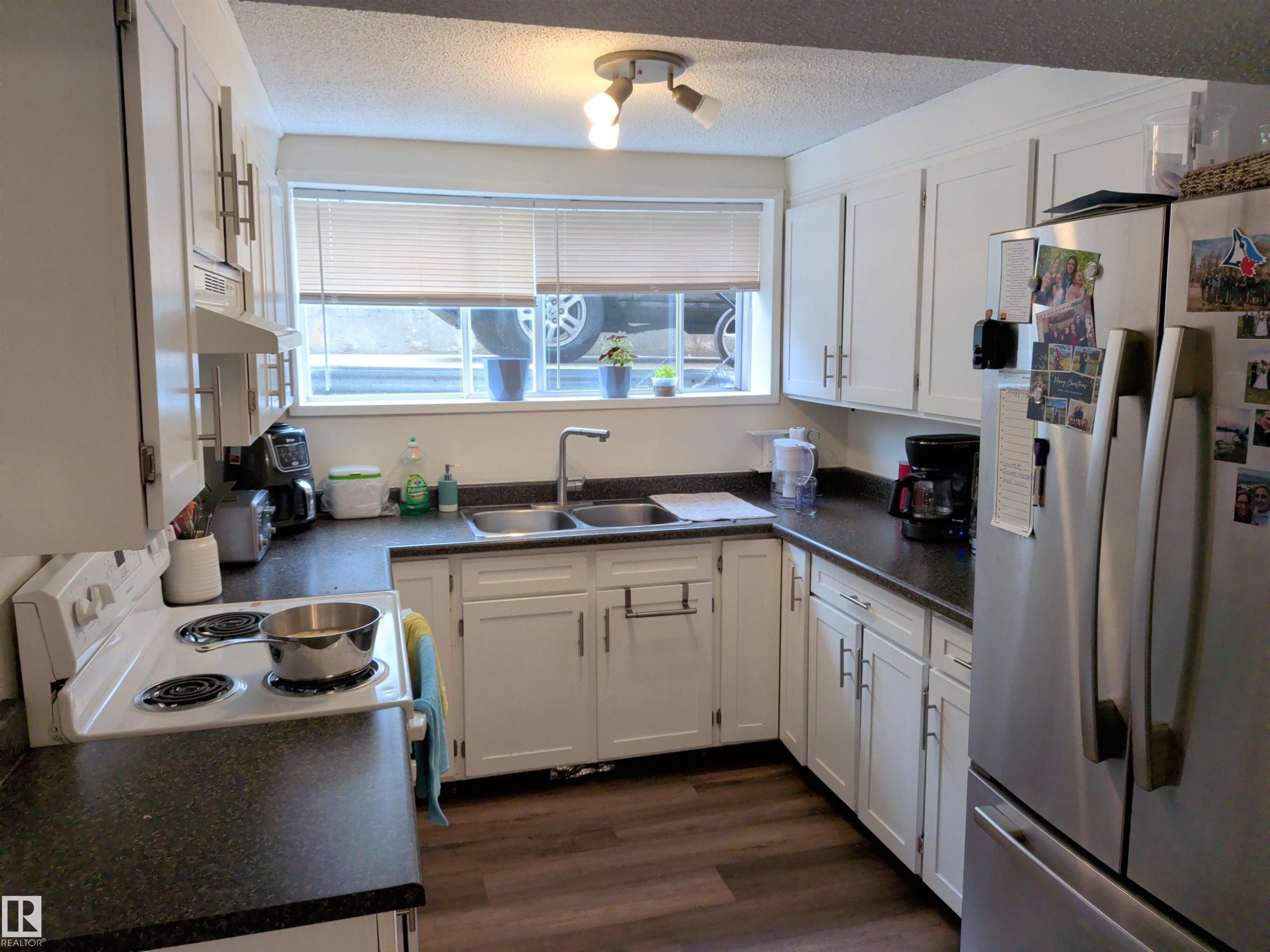 Kitchen featuring white cabinetry, dark countertops, wood-finish flooring, a double basin sink, and a stainless steel refrigerator - 31 Brown Street, Stony Plain, AB