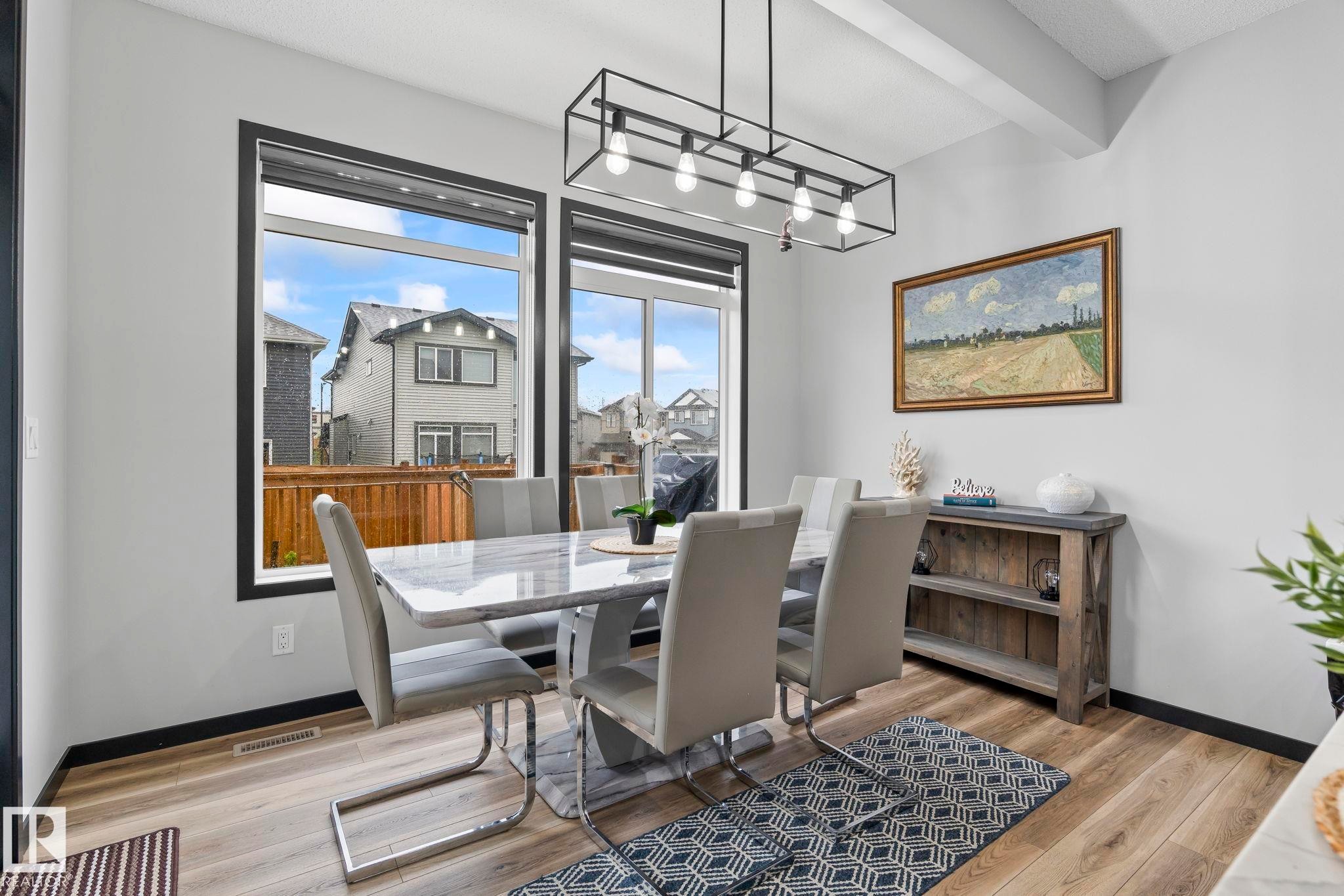 Dining area featuring large windows with dark trim, wood-finish flooring, and a contemporary linear chandelier with exposed bulbs - 5110 23A Ave Sw, Edmonton, AB