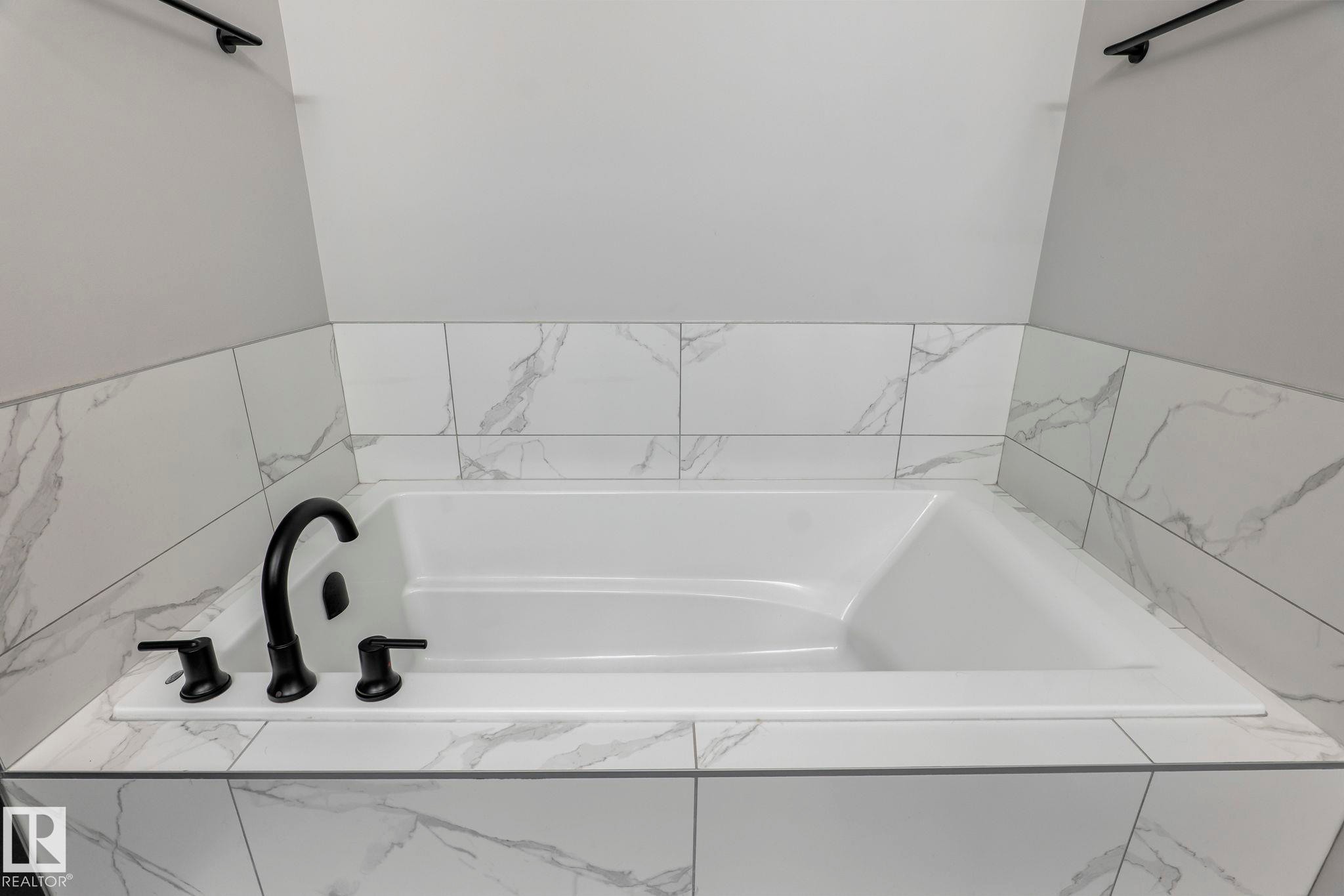 Built-in drop-in bathtub featuring a deck-mounted matte black faucet, surrounded by marbled-patterned tile, and framed by light gray walls - 5110 23A Ave Sw, Edmonton, AB
