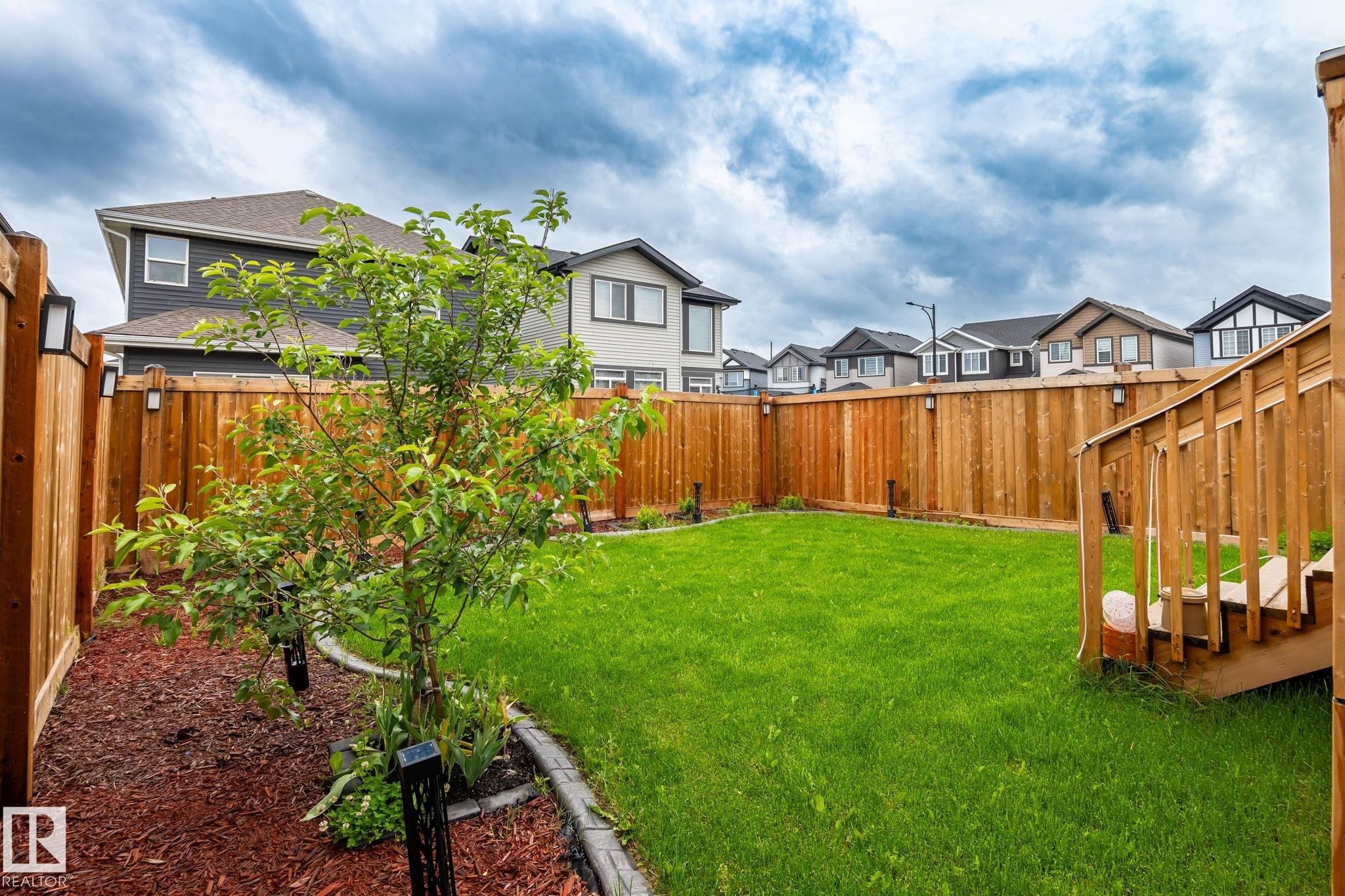 Expansive backyard featuring a manicured lawn, a full-height wooden privacy fence, a wooden deck with stairs, a planted tree, and a mulched garden bed with decorative edging - 5110 23A Ave Sw, Edmonton, AB