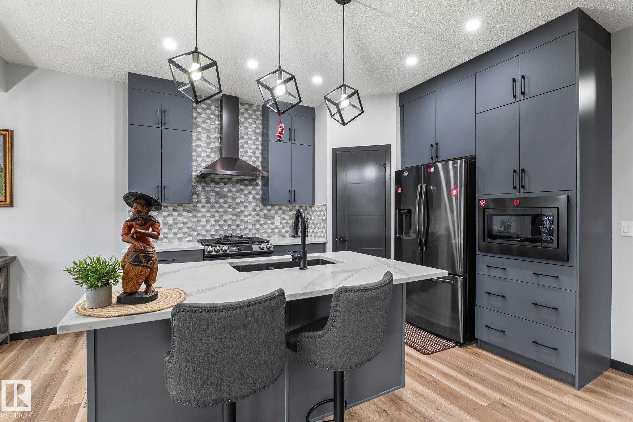 Modern kitchen featuring gray flat-panel cabinetry, a central island with a light-toned countertop, stainless steel appliances, and a mosaic tile backsplash - 5110 23A Ave Sw, Edmonton, AB