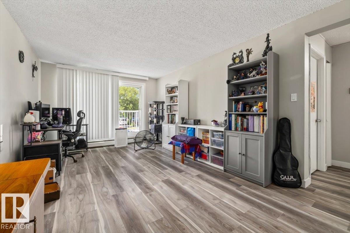 Spacious room featuring wood-finish flooring, a large window with vertical blinds, and neutral wall tones - #202 10330 113 St Nw, Edmonton, AB