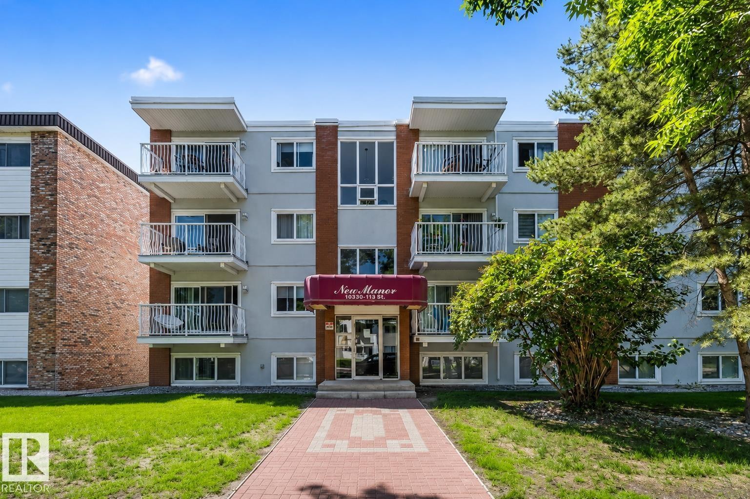Multi-story building featuring light grey facade, individual balconies with white railings, and a red entrance awning - #202 10330 113 St Nw, Edmonton, AB