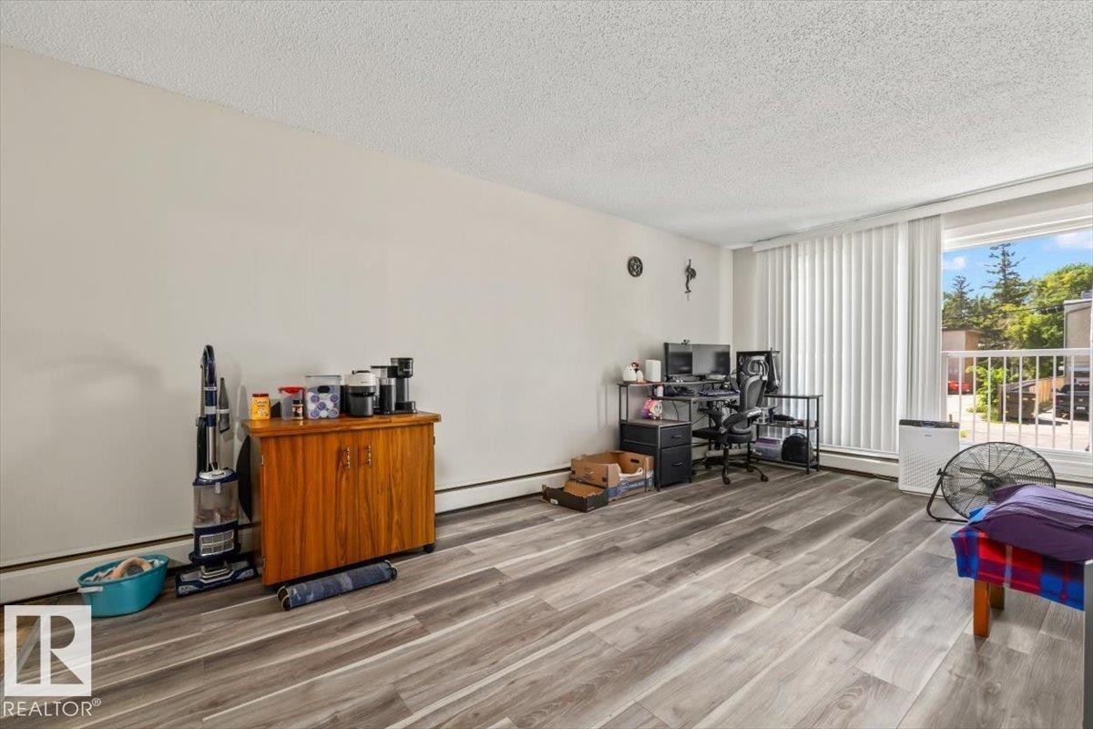 Spacious room featuring wood-finish flooring, white walls, and a large window with vertical blinds - #202 10330 113 St Nw, Edmonton, AB