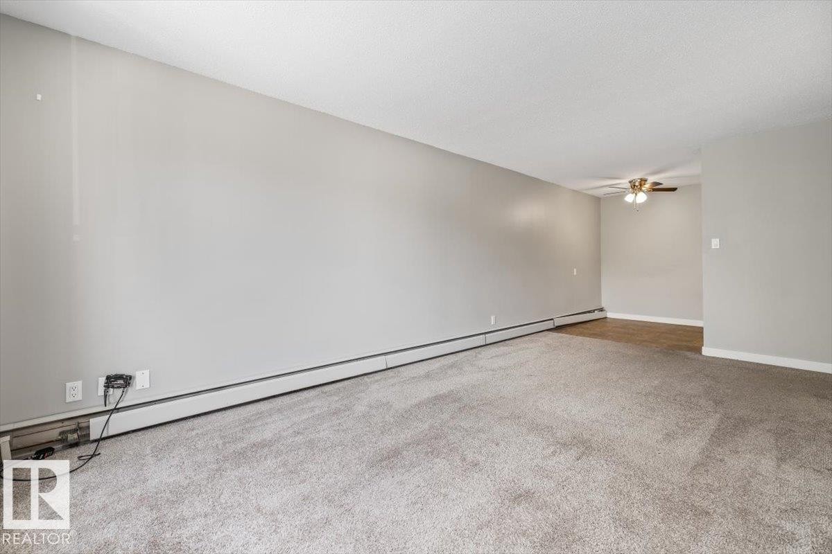 Spacious room featuring neutral wall tones, textured carpeting, baseboard heating, and a ceiling fan with integrated lighting - #306 10330 113 St Nw, Edmonton, AB