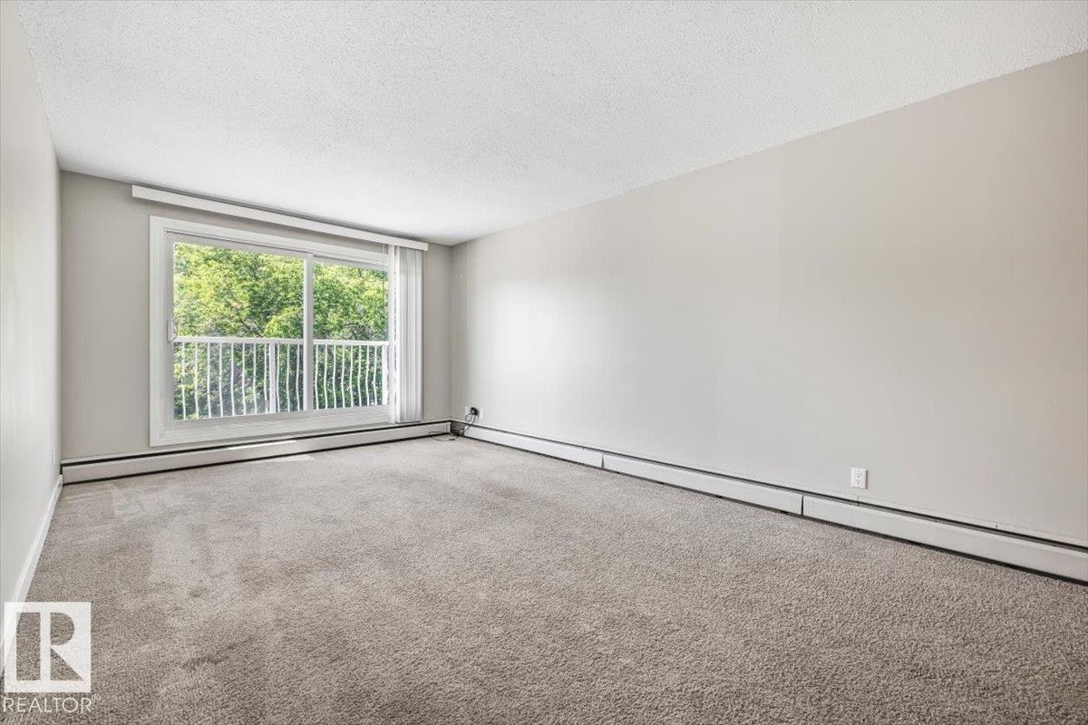 Spacious room featuring light gray carpeting, neutral wall paint, and white baseboard heating - #306 10330 113 St Nw, Edmonton, AB